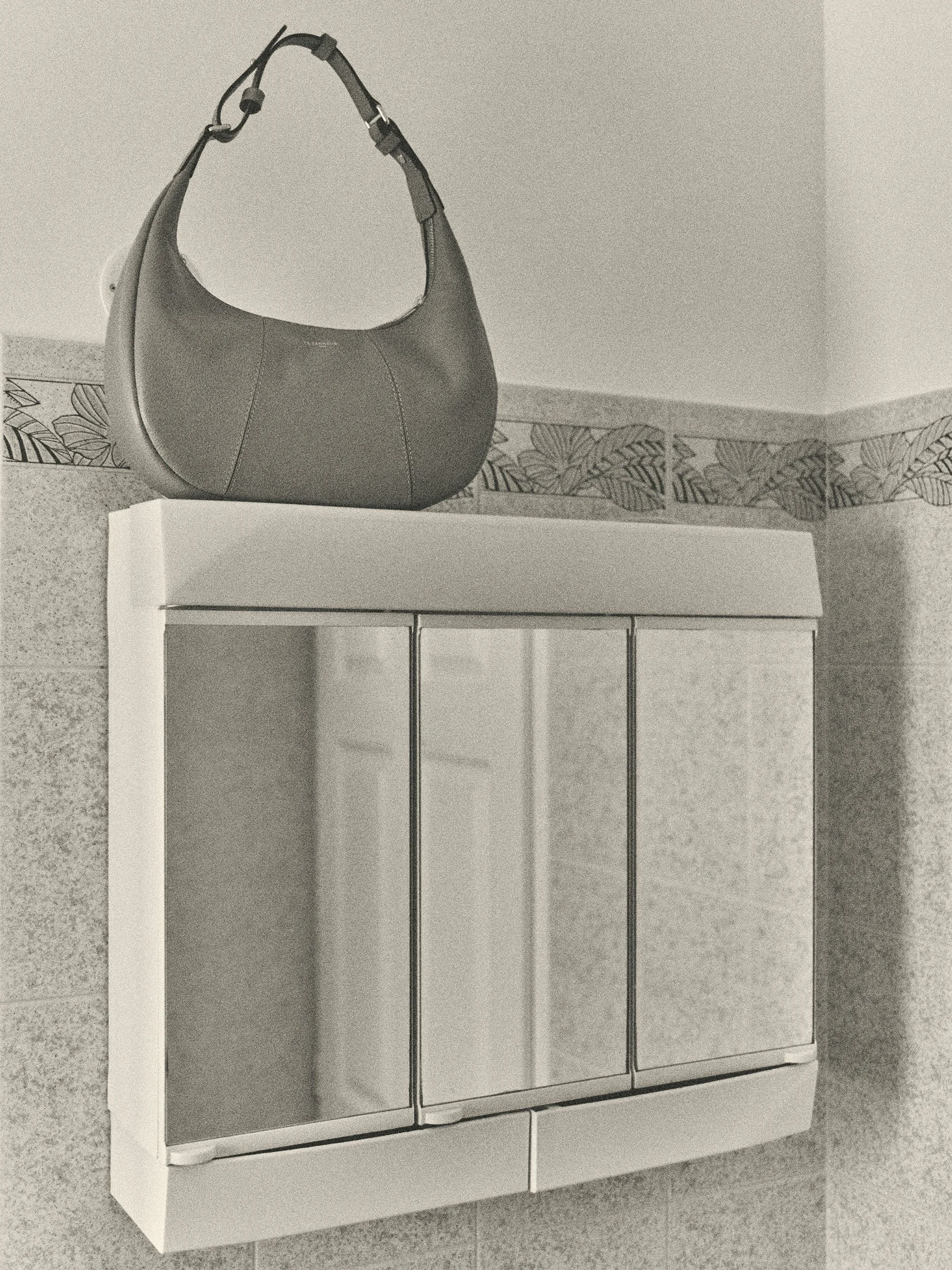 Editorial fashion still life of a Le Tanneur leather hobo bag displayed on a vintage bathroom cabinet, photographed in black and white.