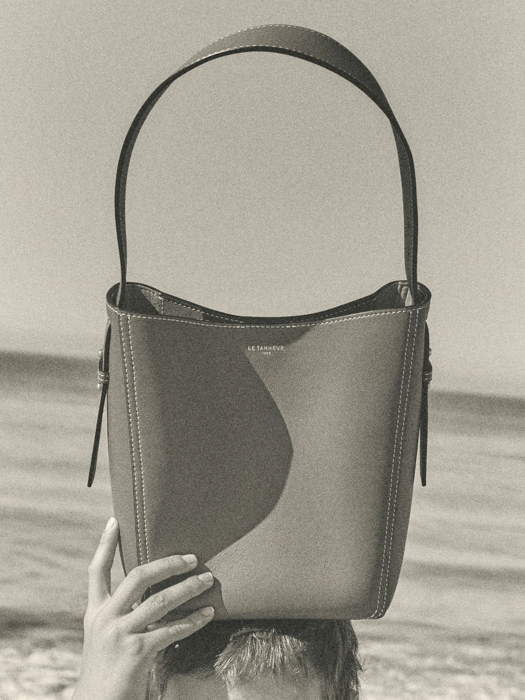 Editorial fashion portrait of a brown Le Tanneur leather shoulder bag held in front of the face on a beach, photographed in black and white.
