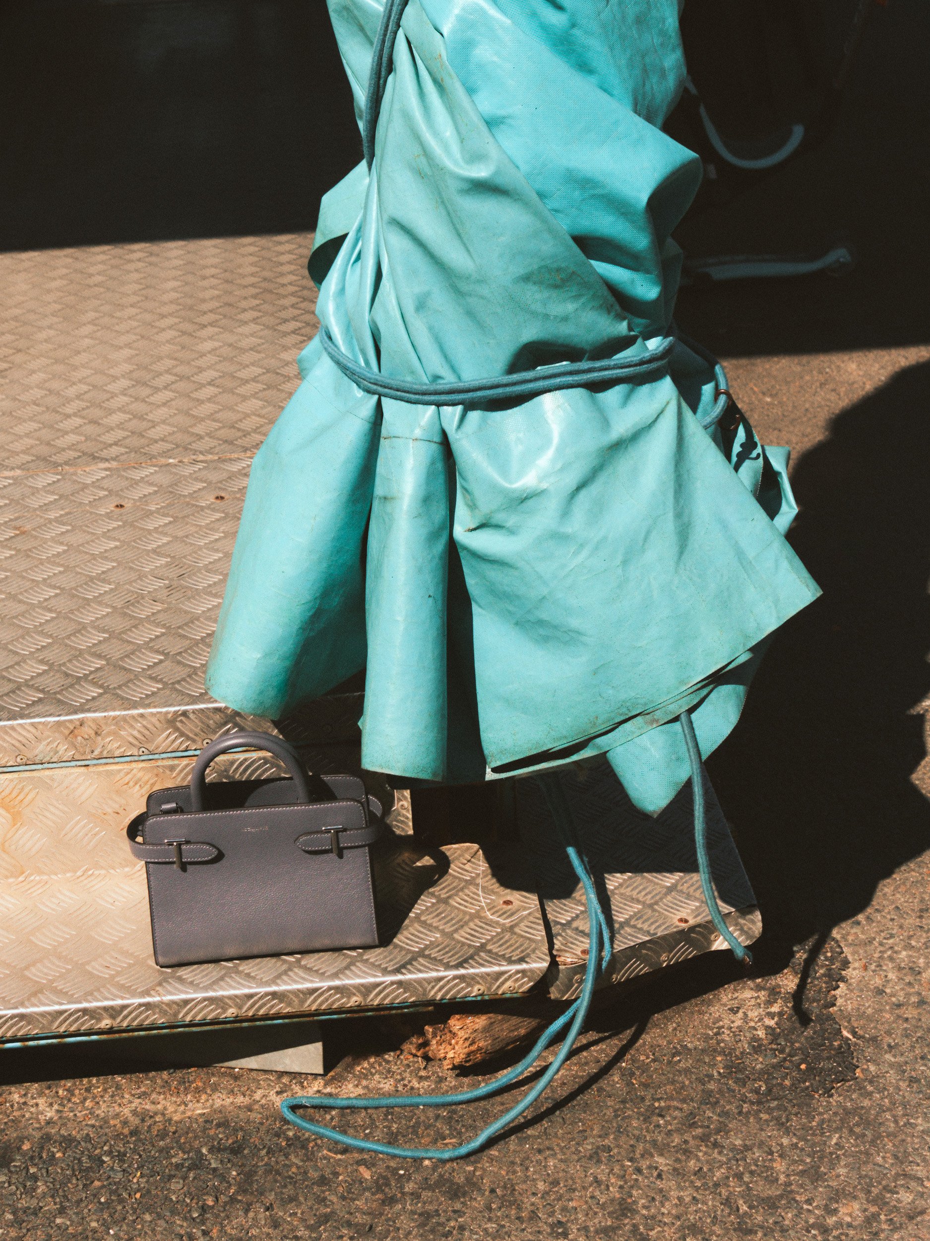 Editorial fashion still life of a black Le Tanneur leather handbag beside a turquoise fairground cover, photographed in strong sunlight and shadow.