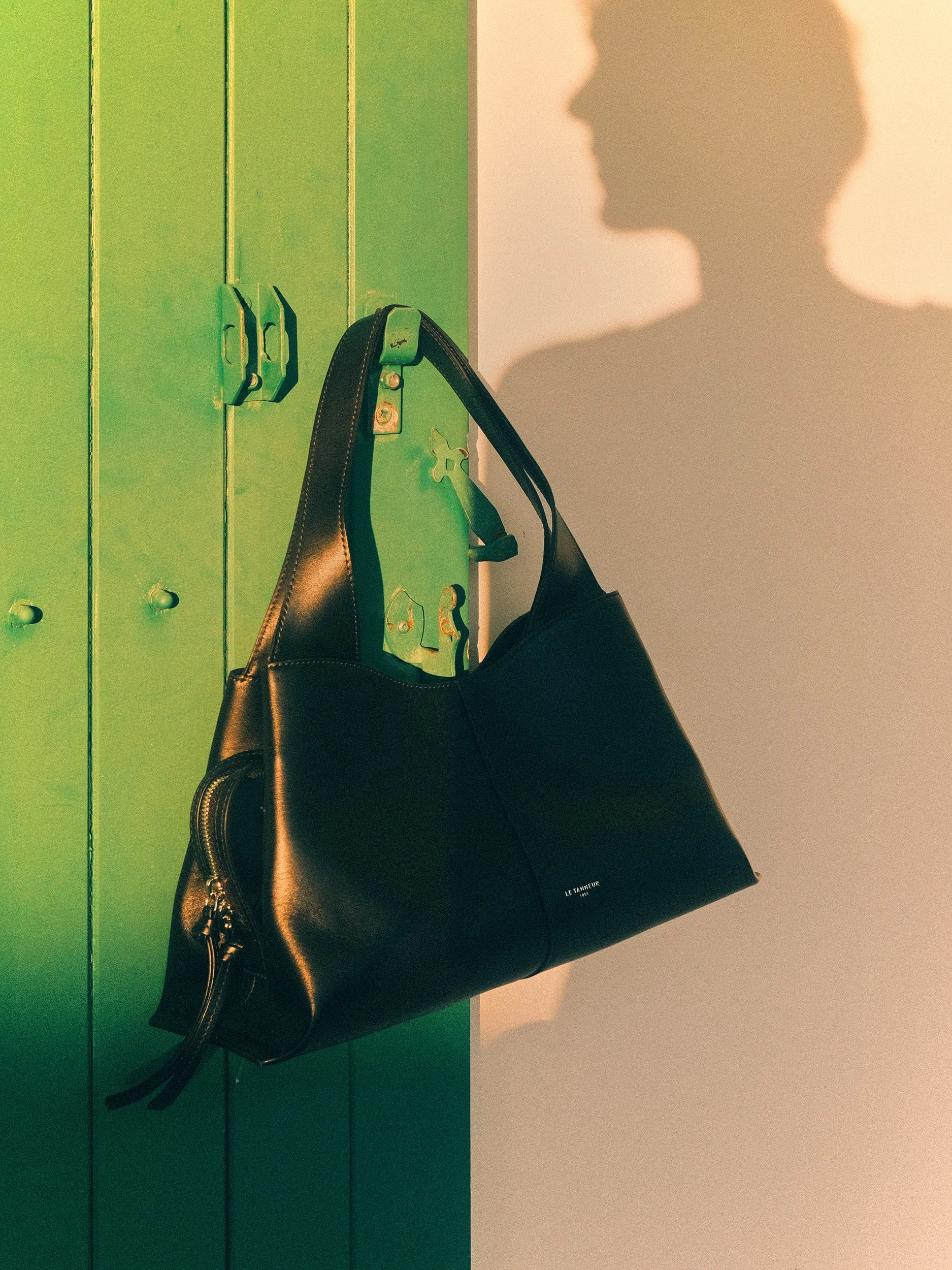 Editorial fashion still life of a black Le Tanneur leather handbag hanging from a green metal door with a woman’s shadow cast on the wall in warm sunlight.