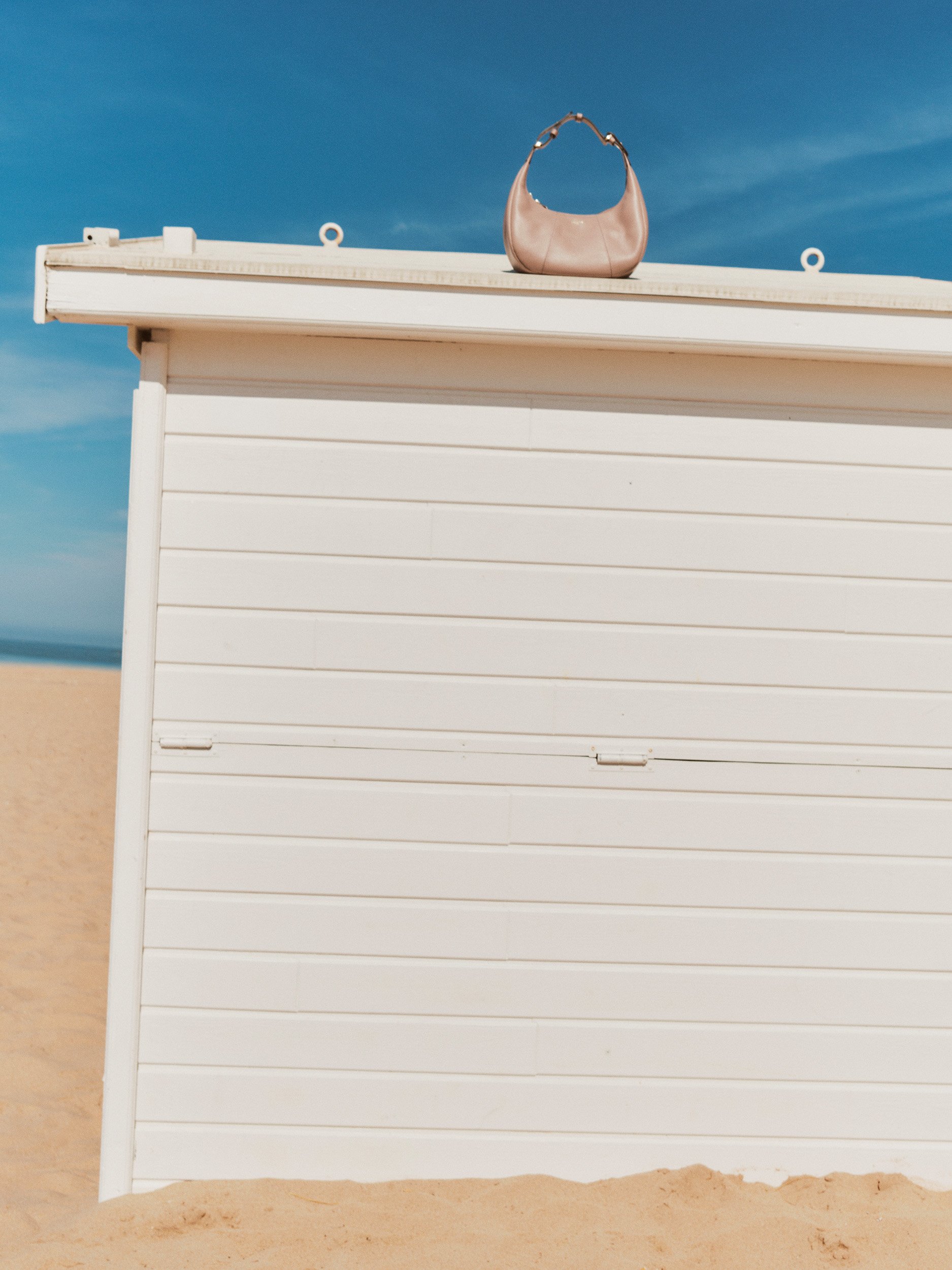 Editorial fashion still life of a taupe Le Tanneur leather hobo bag displayed on a white beach hut against a bright blue sky.
