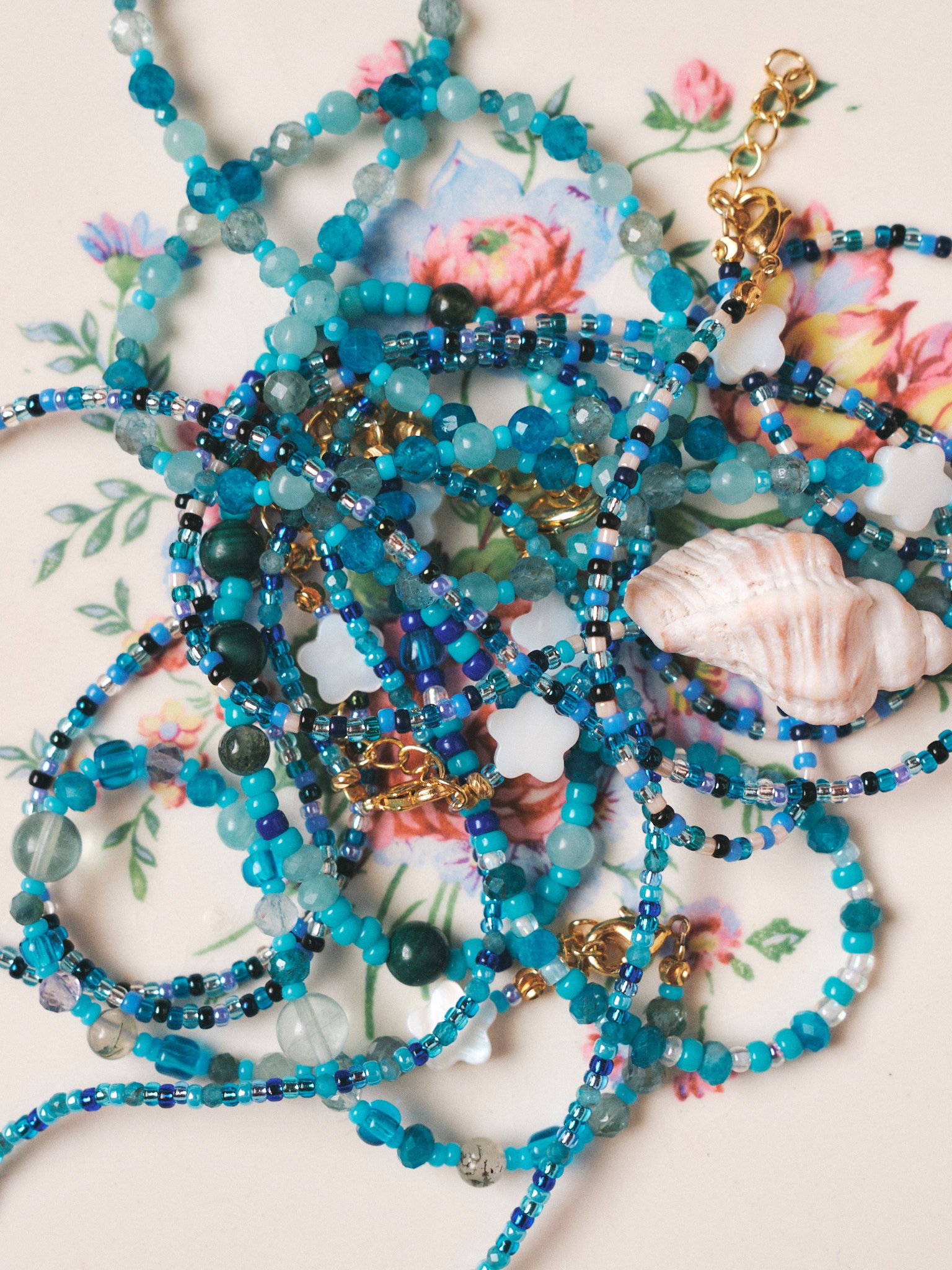 Editorial jewelry still life of turquoise and blue beaded necklaces with a seashell on a vintage floral plate, photographed in soft natural light.