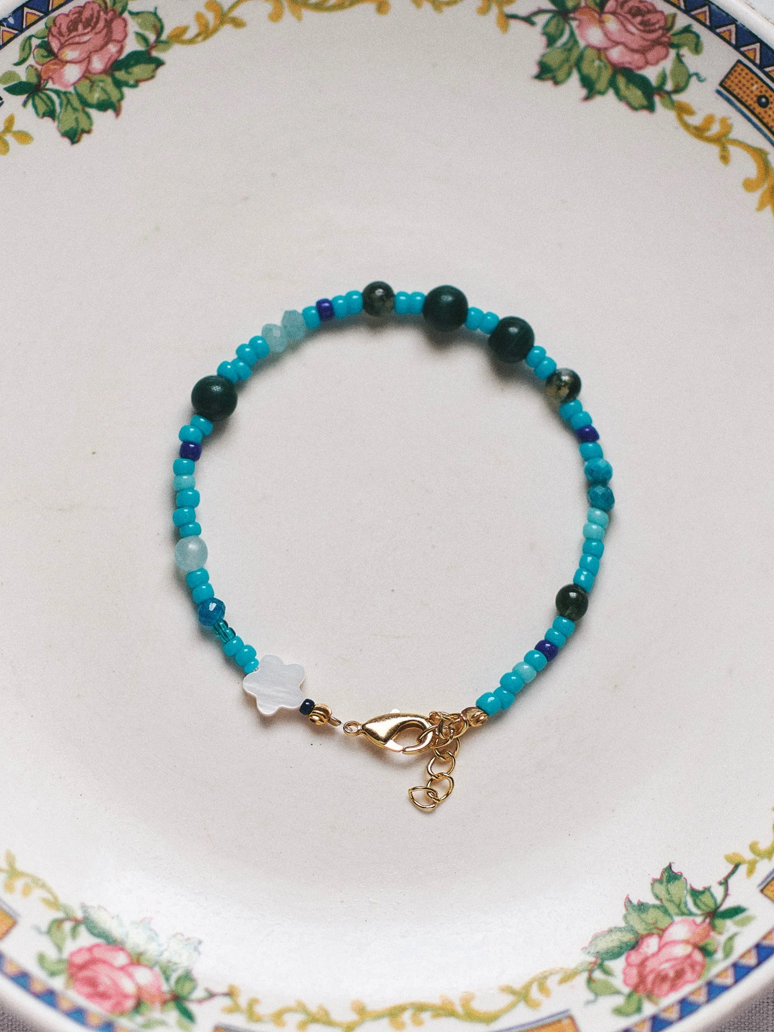 Editorial jewelry still life of a turquoise and dark green beaded bracelet on a vintage floral plate, photographed in soft natural light.