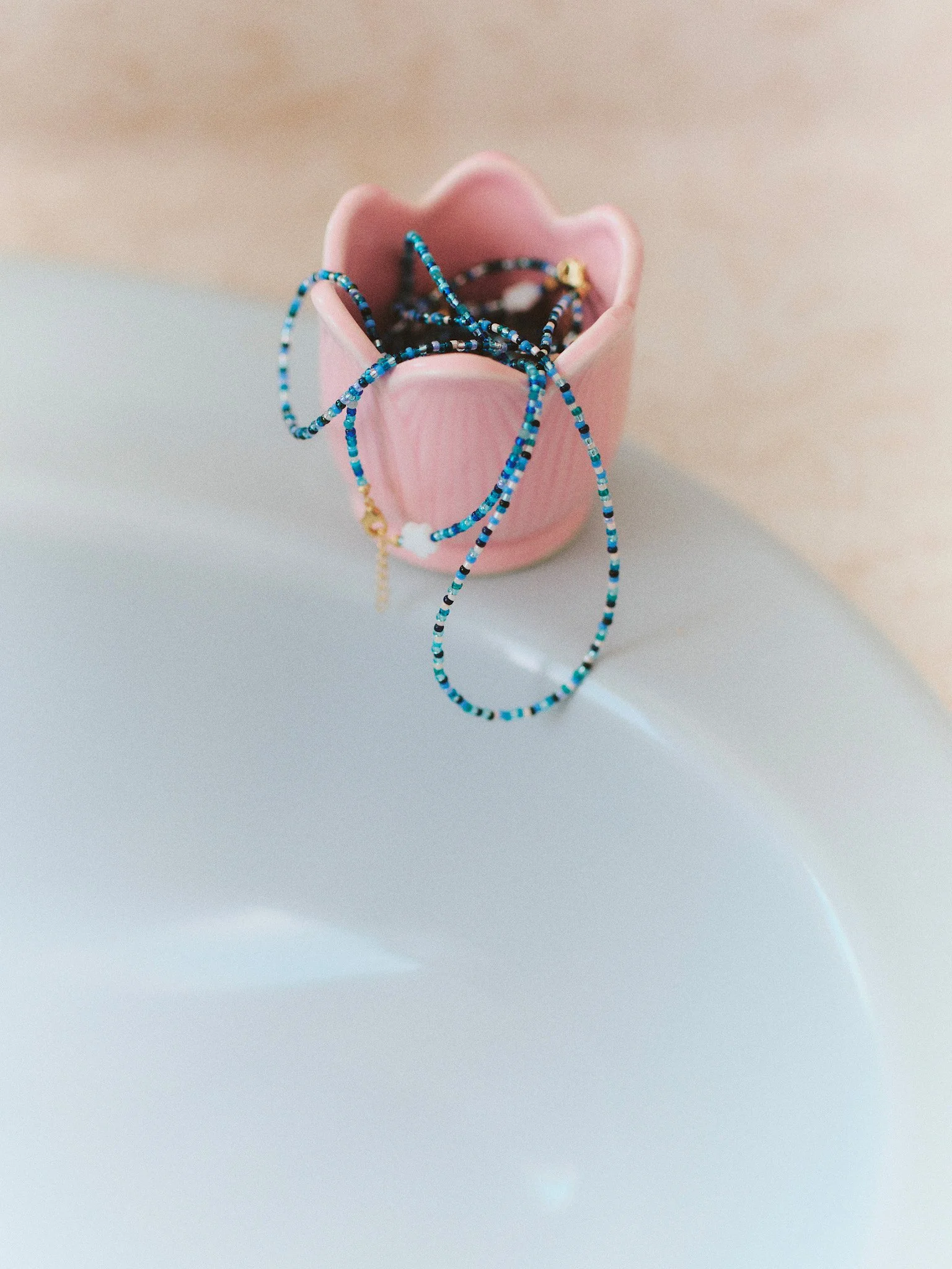 Editorial jewelry still life of delicate blue beaded necklaces draped from a pink ceramic cup, photographed in soft natural light.