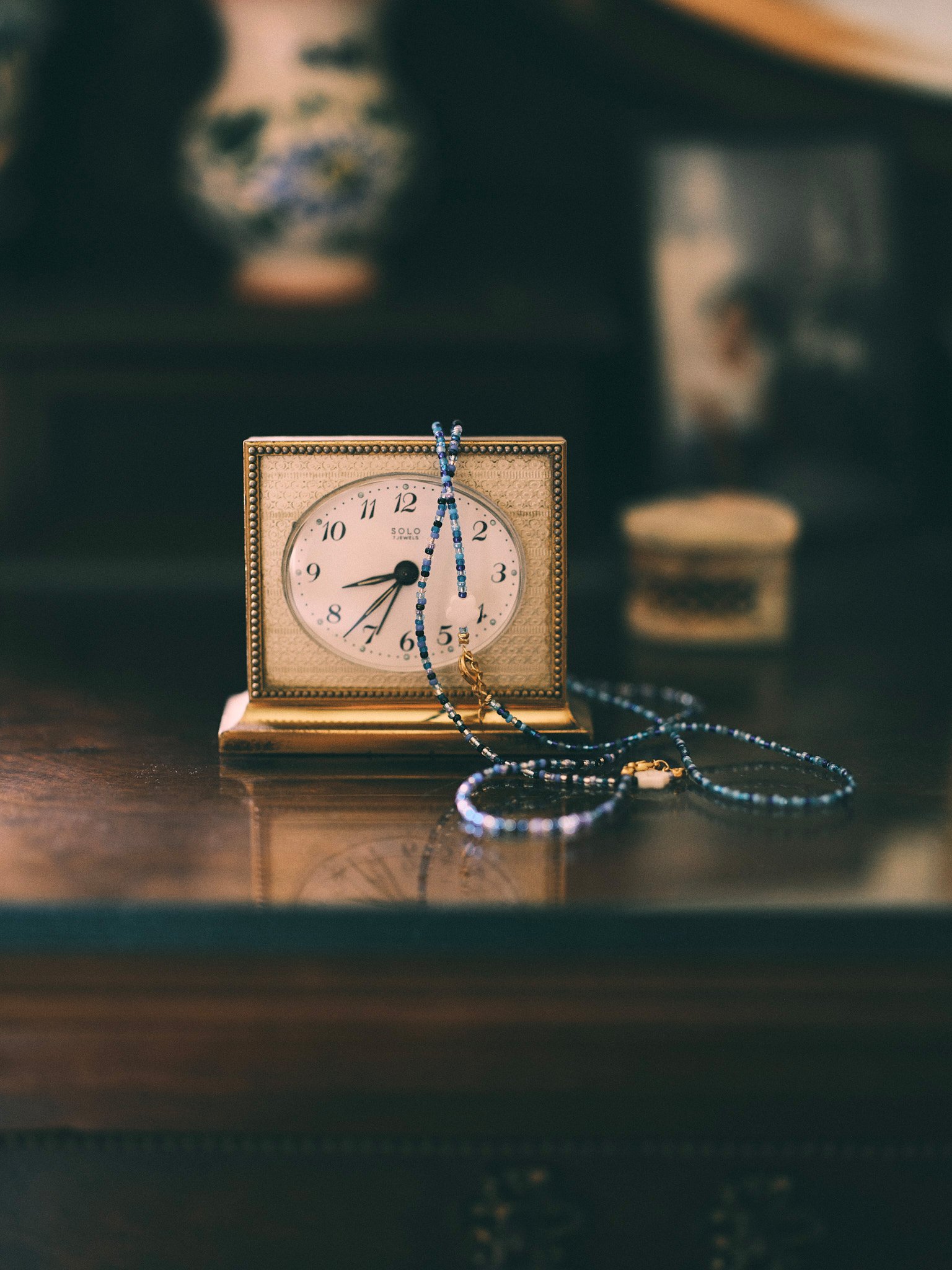 Editorial jewelry still life of delicate blue beaded necklaces draped over a vintage brass clock in a softly lit interior.