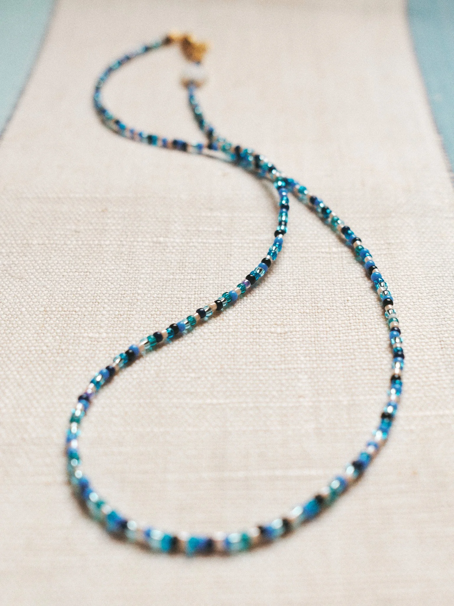 Editorial jewelry still life of a delicate blue beaded necklace arranged on textured white linen, photographed in soft natural light.