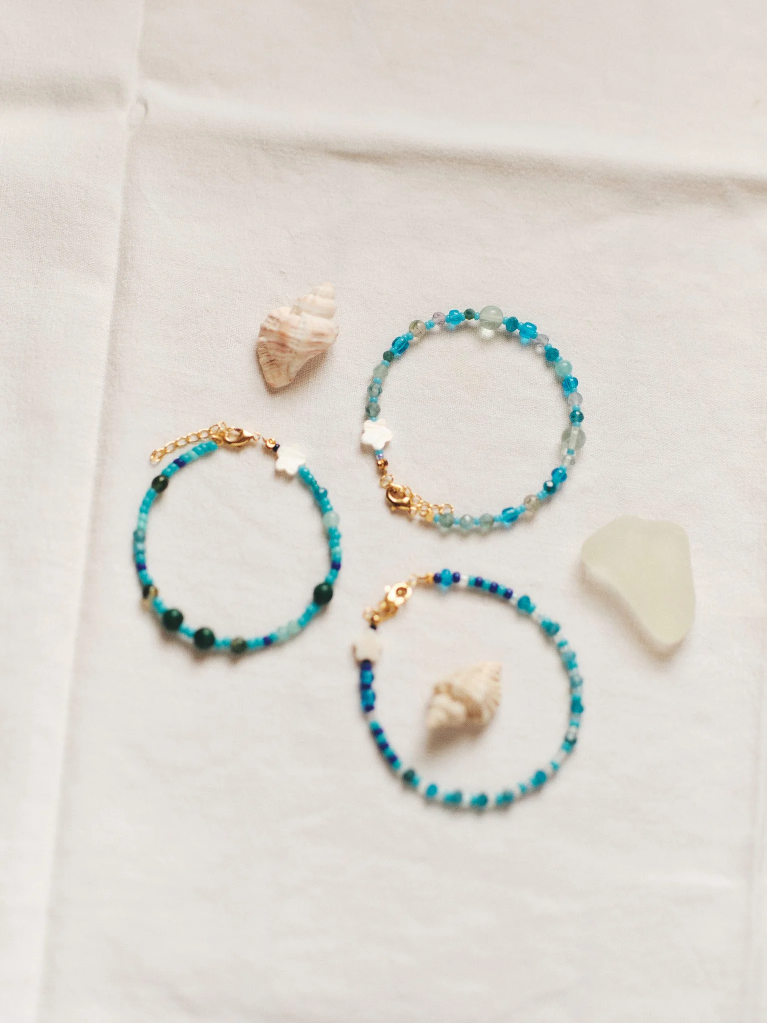 Editorial jewelry still life of three blue and turquoise beaded bracelets with seashells on white linen, photographed in soft natural light.