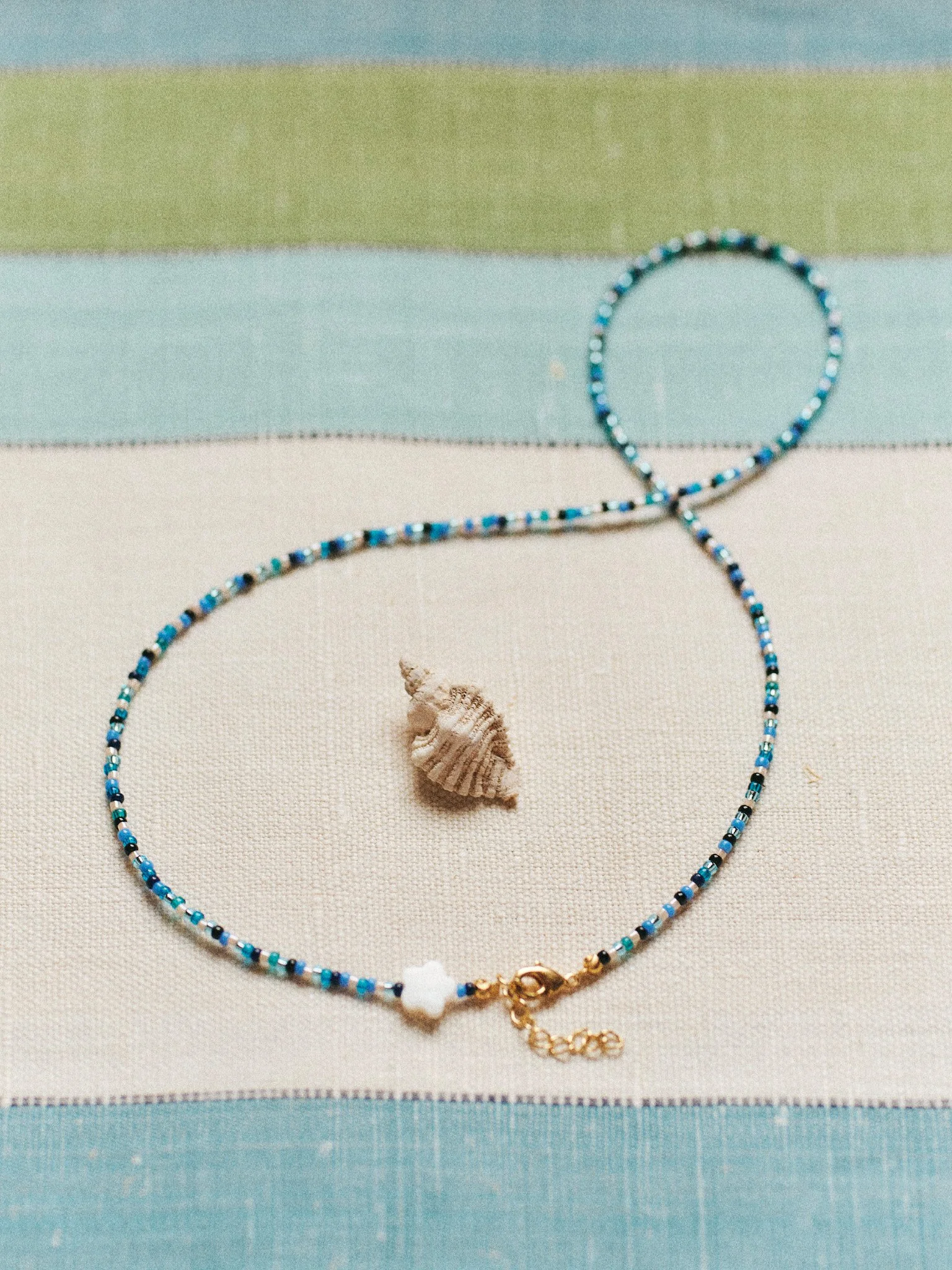 Editorial jewelry still life of a delicate blue beaded necklace with a small seashell on striped linen, photographed in soft natural light.