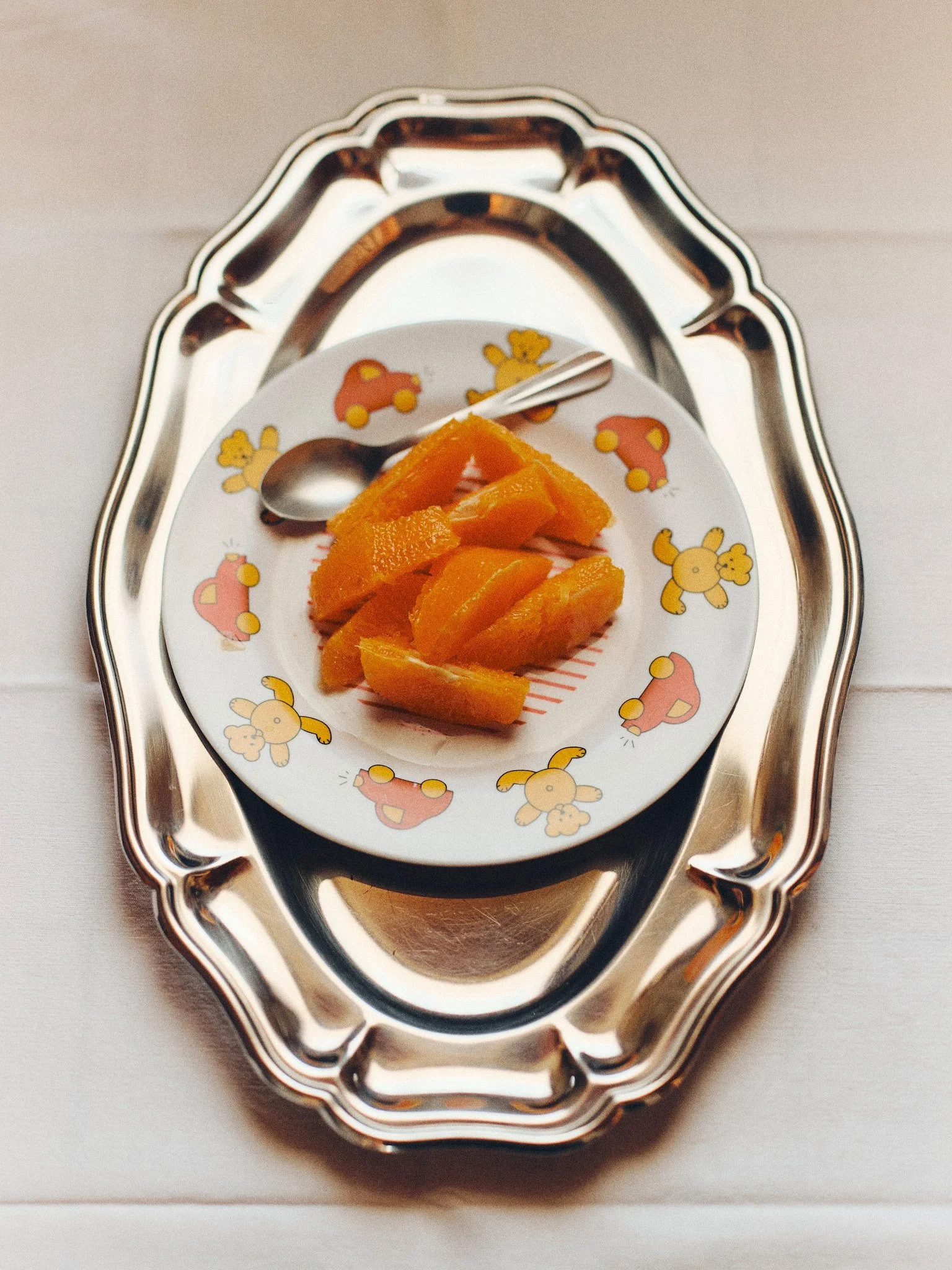 Editorial food and tableware still life of orange slices on a vintage children’s plate arranged on a silver serving tray in soft natural light.