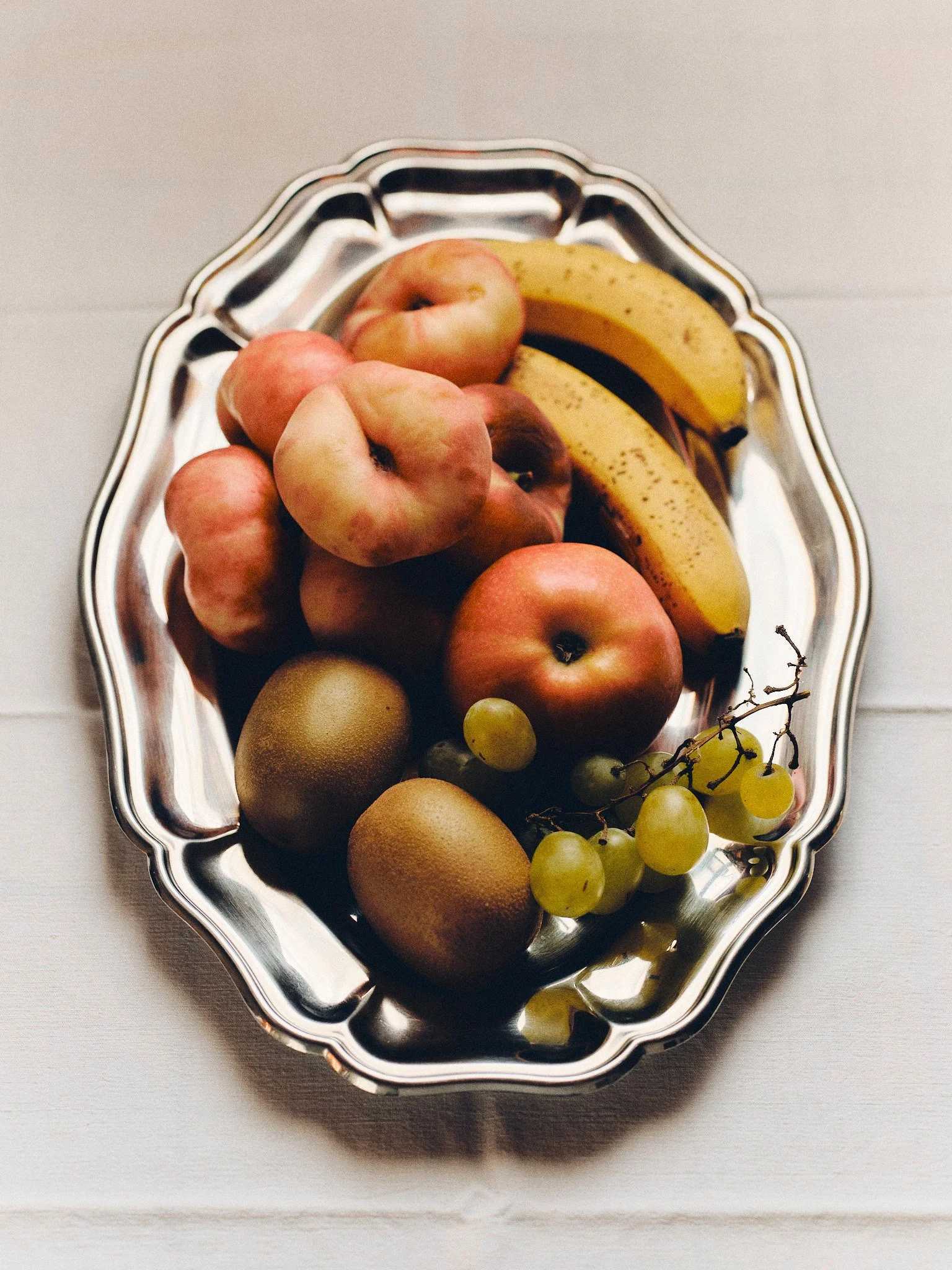 Editorial food still life of peaches, bananas, apples, kiwis and grapes arranged on a vintage silver serving tray in soft natural light.