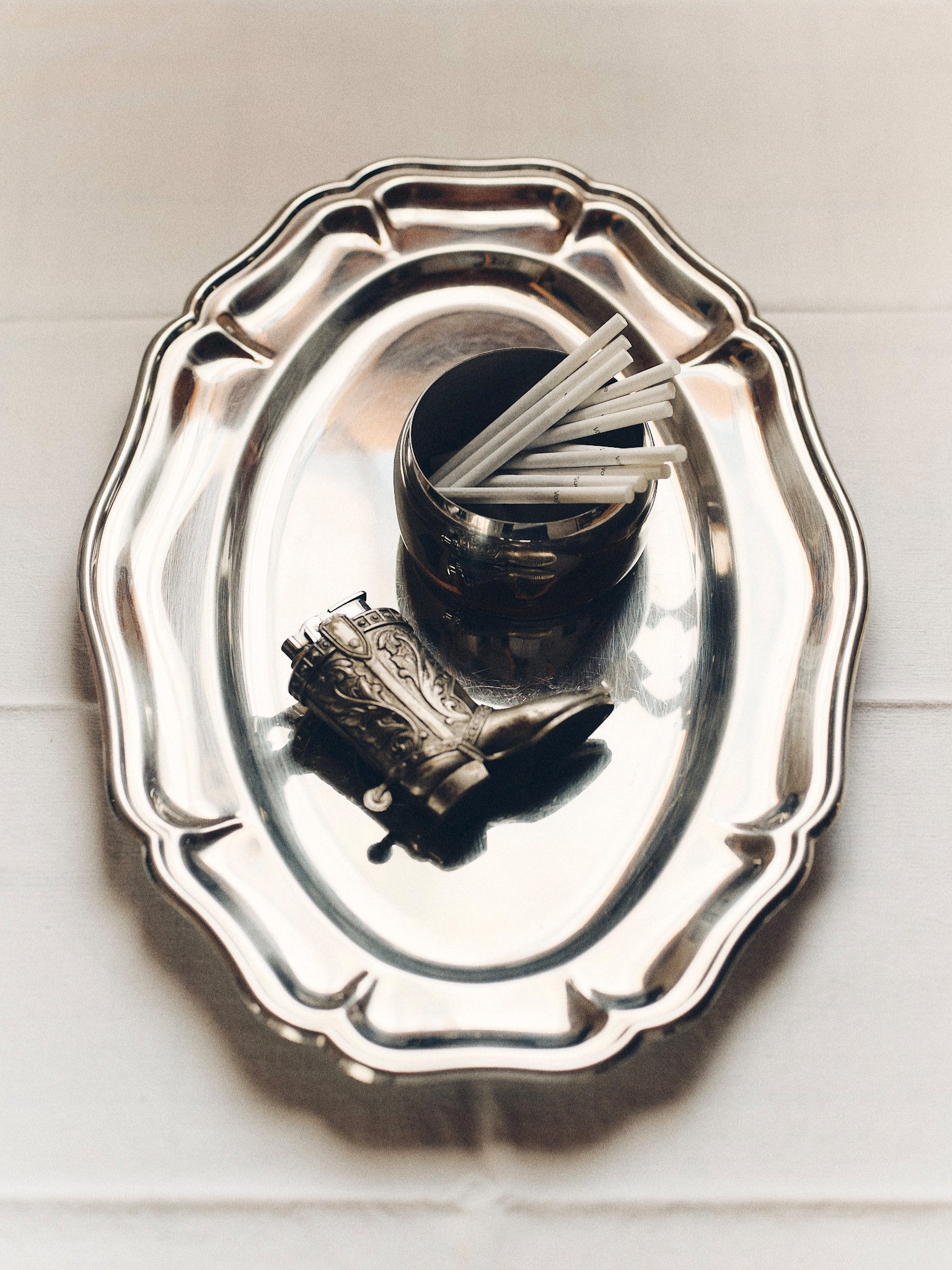 Editorial still life of vintage smoking accessories with white cigarettes and a sculptural metal object arranged on a silver serving tray.