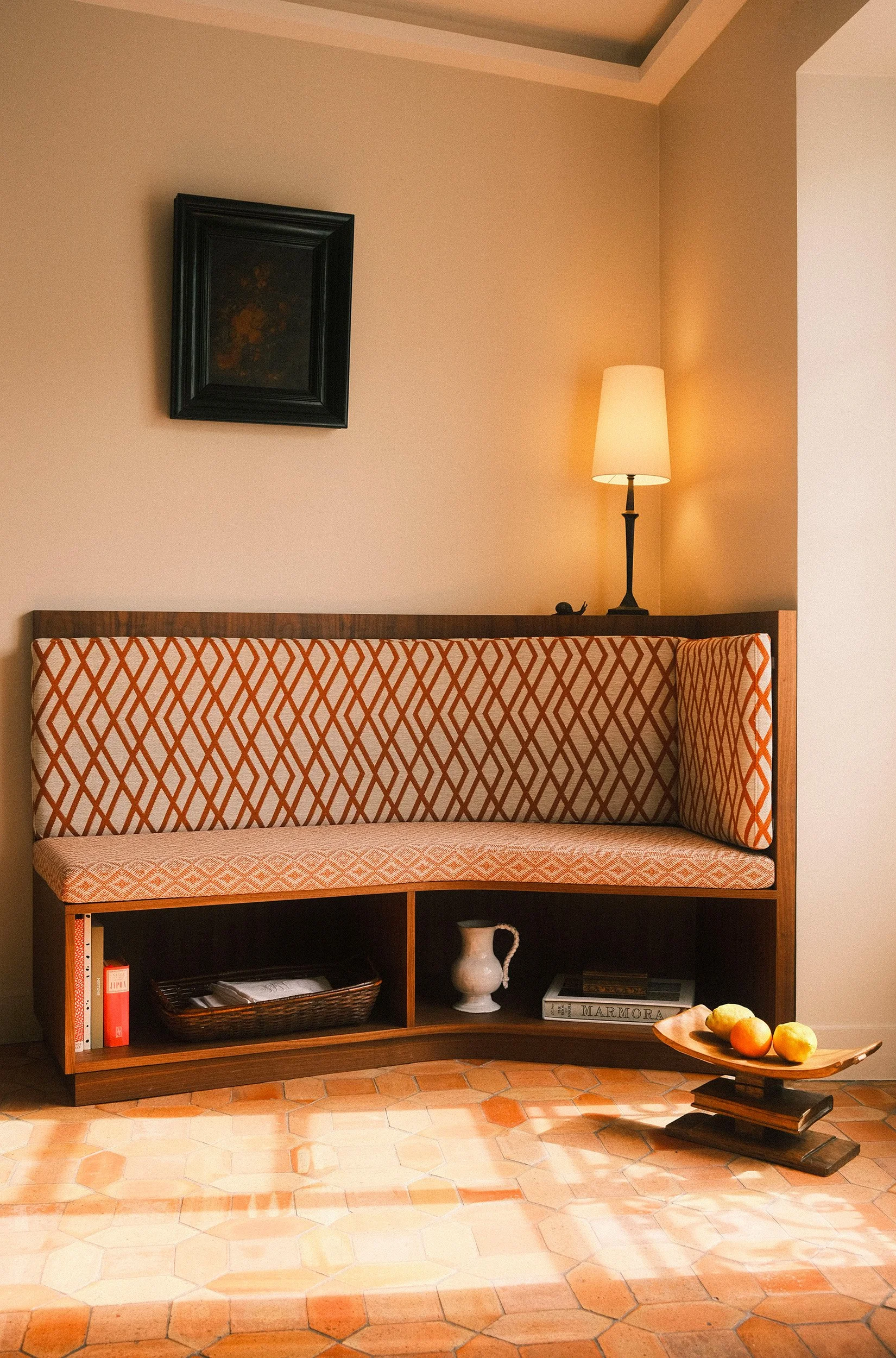 Editorial interior photography of a warm sunlit living space with custom wooden seating, patterned upholstery, terracotta floor tiles and sculptural decor.