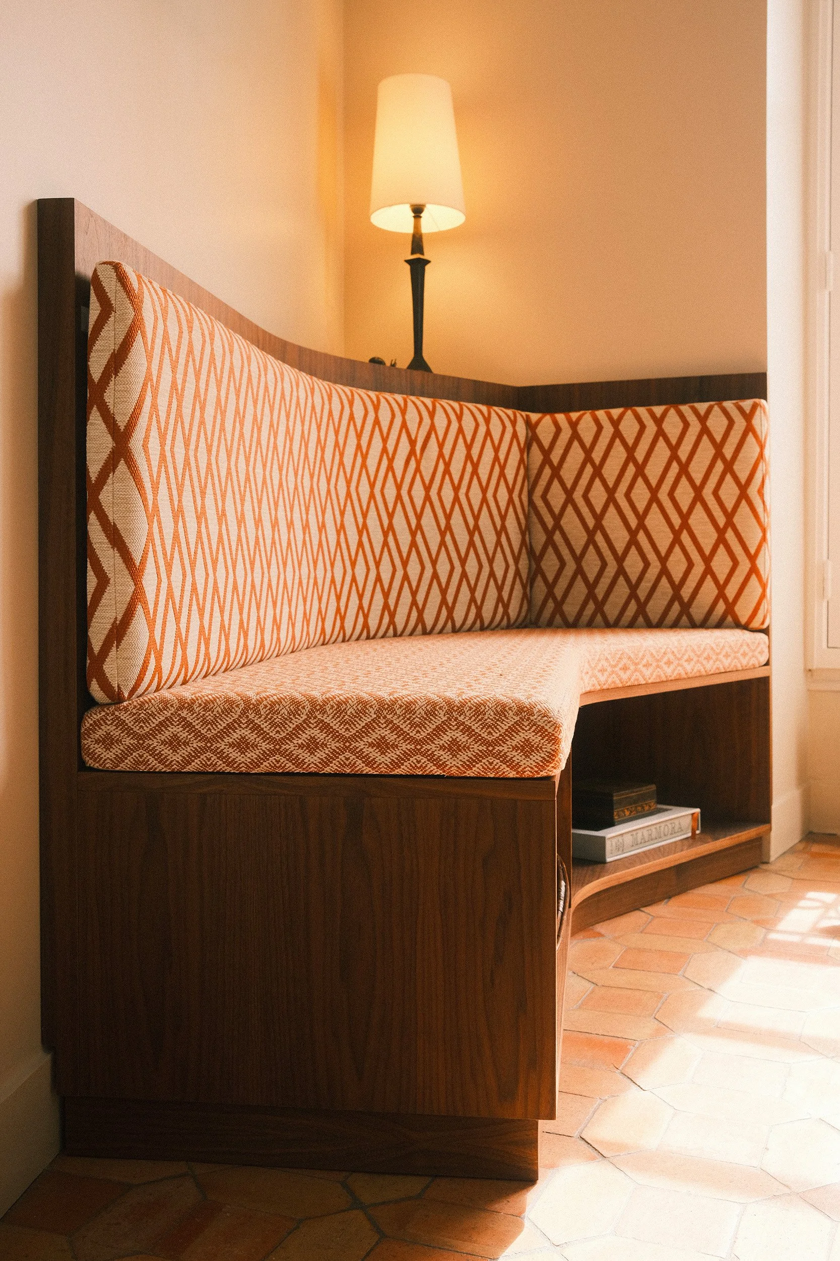 Editorial interior photography of a custom dark wood corner banquette with orange geometric upholstery, terracotta floor tiles and warm ambient light.