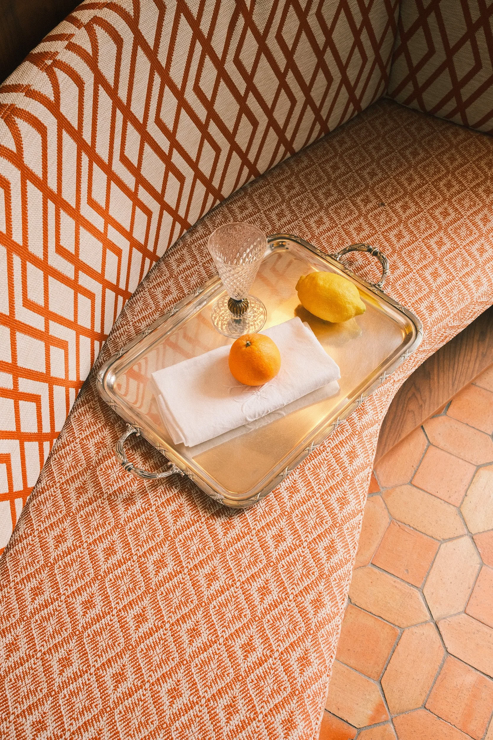 Editorial interior and still life photography of a silver tray with citrus fruit and crystal glass on a patterned upholstered bench, photographed in warm natural light.