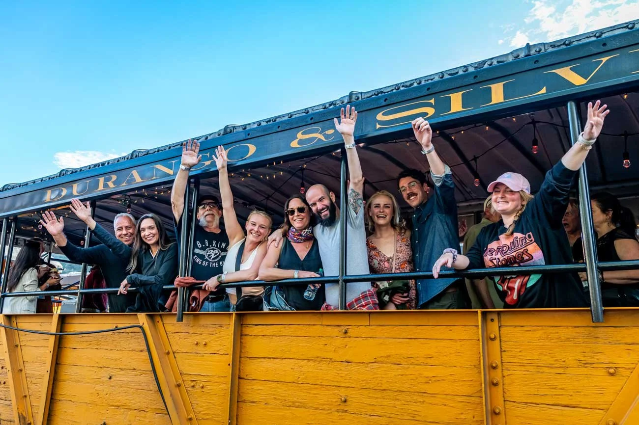 Passengers smile and raise drinks while riding in the open-air gondola aboard the Durango Blues Train, enjoying live blues music, fresh mountain air, and scenic views through the San Juan Mountains.