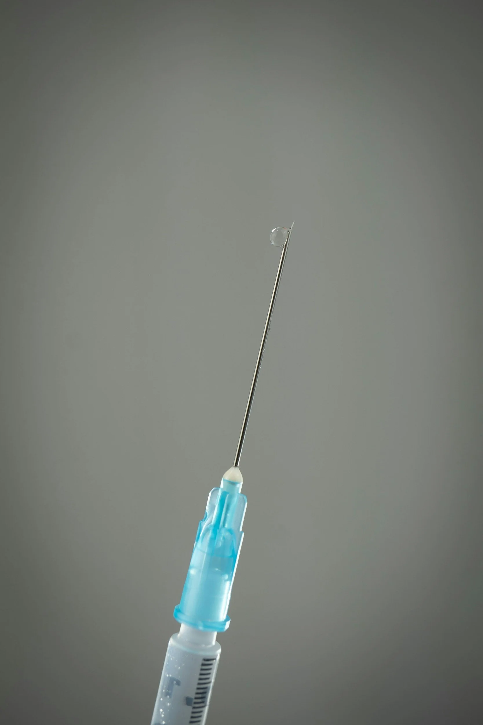A close-up of a medical syringe with a needle and a small droplet of liquid at the tip, against a plain gray background.