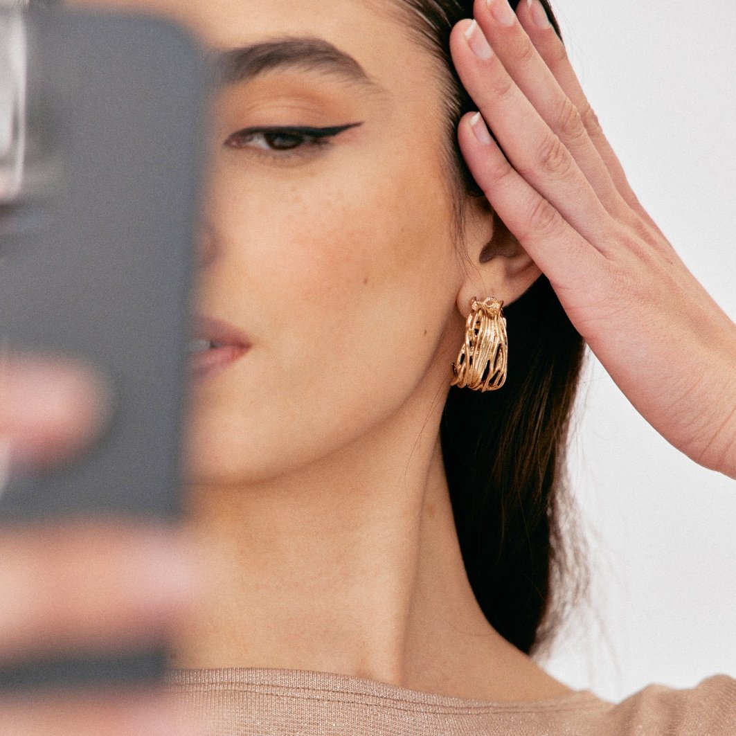 Close-up of a woman with dark hair and gold hoop earrings taking a selfie, with her face partially visible and one hand touching her hair.