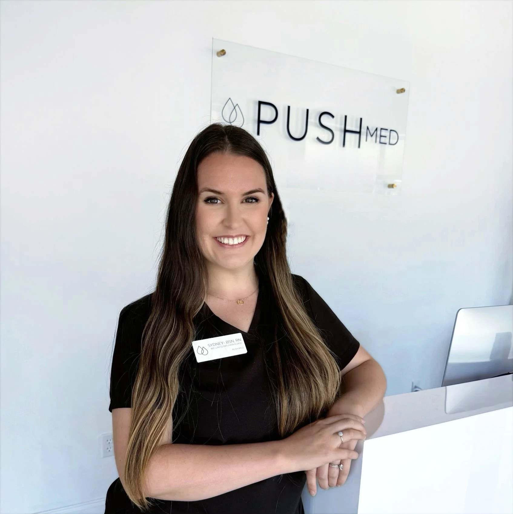 A smiling woman with long brown hair standing at a clinic reception desk. She is wearing a black uniform with a name tag and is positioned in front of a sign that says 'PUSH MED' on a white wall.