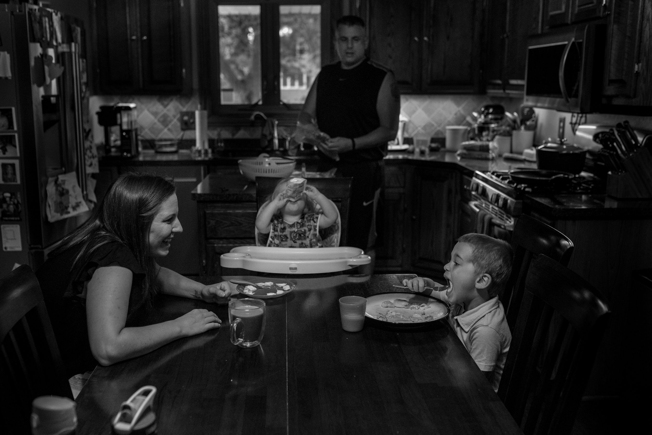 Family of four eating breakfast. Mom smiling at son eating eggs, daughter eating a pouch and dad drying dishes