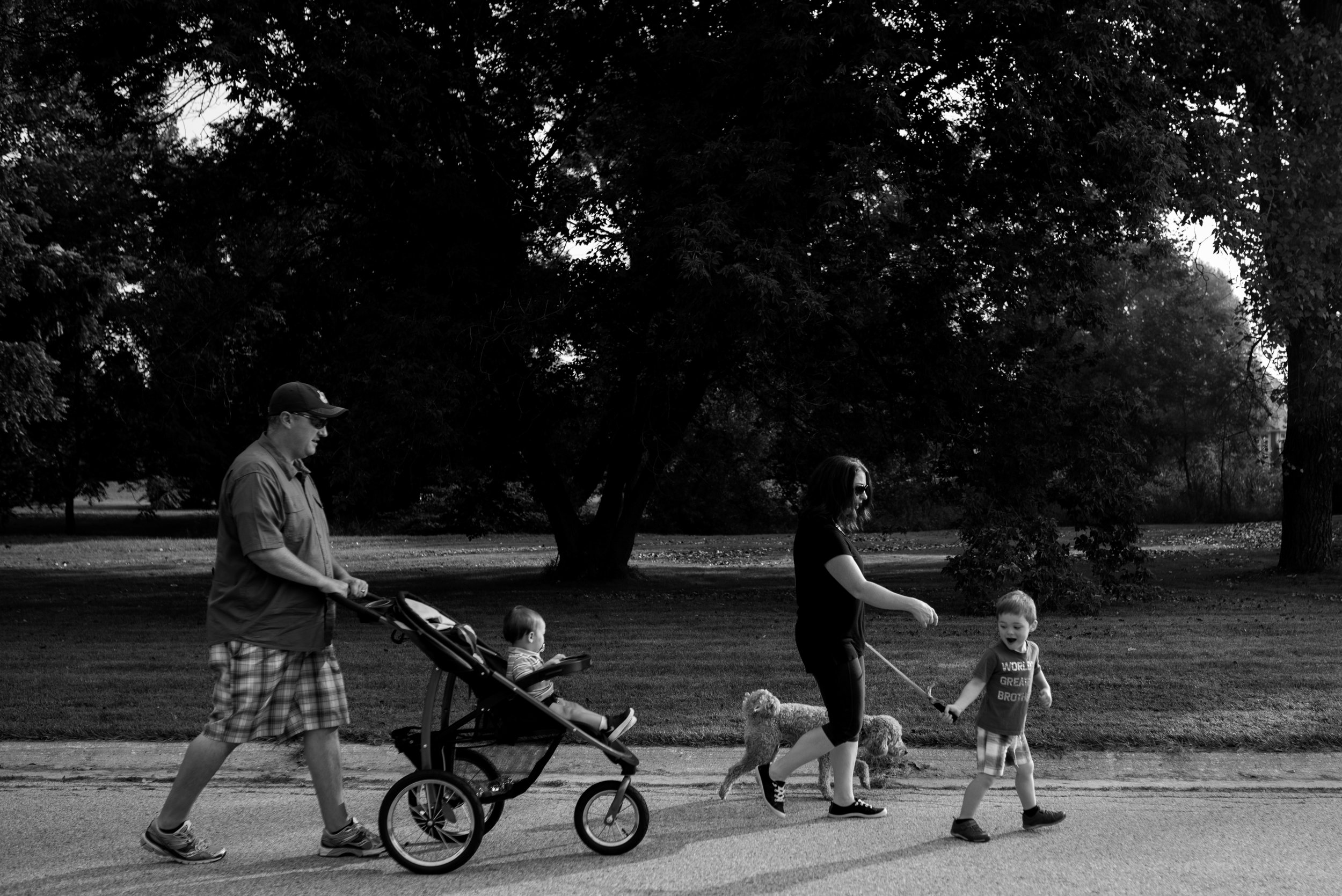 Family walking down the road. Dad pushing stroller with toddler boy, mom and son walking dog