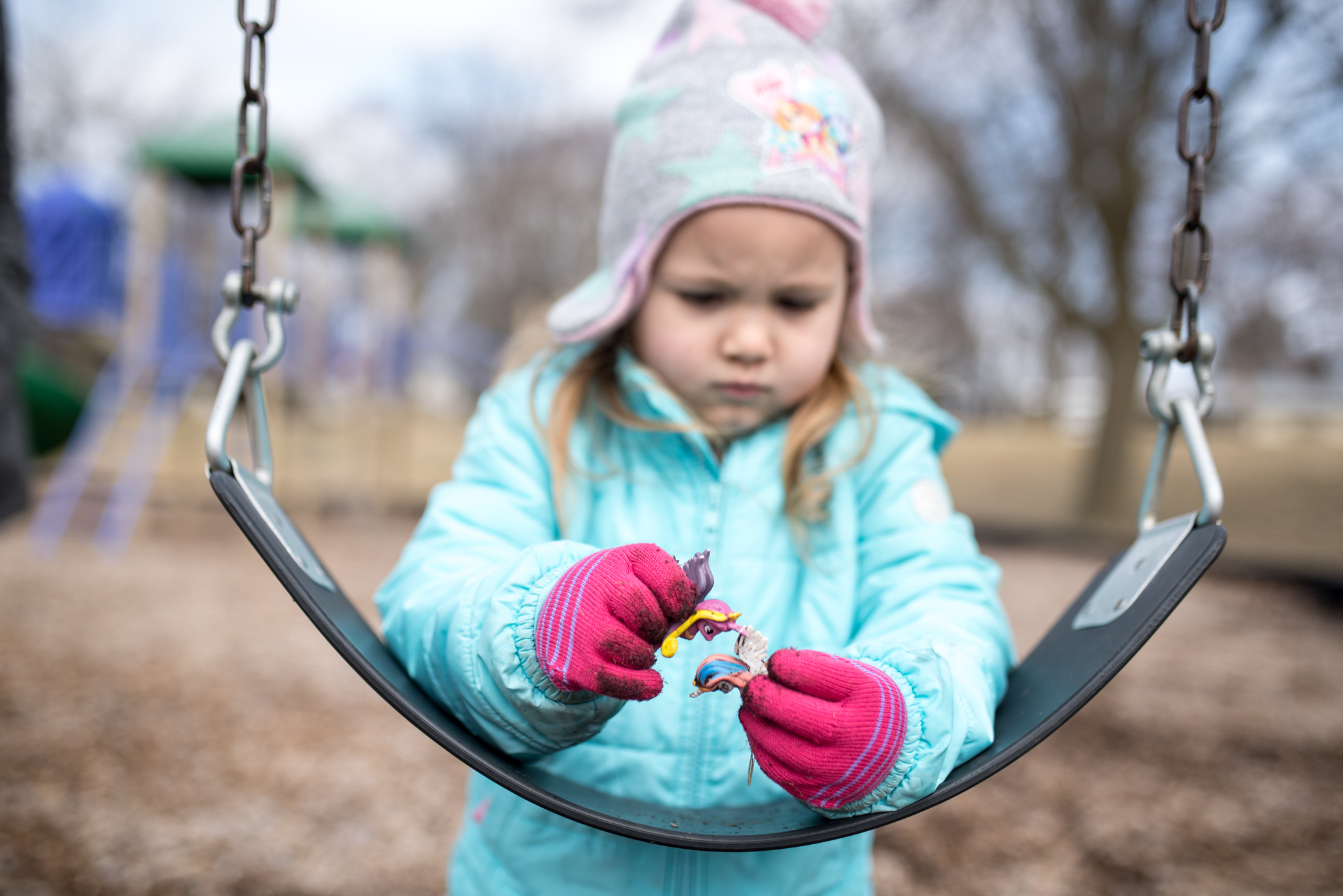 Young girl playing with pony toys on a swing at a park