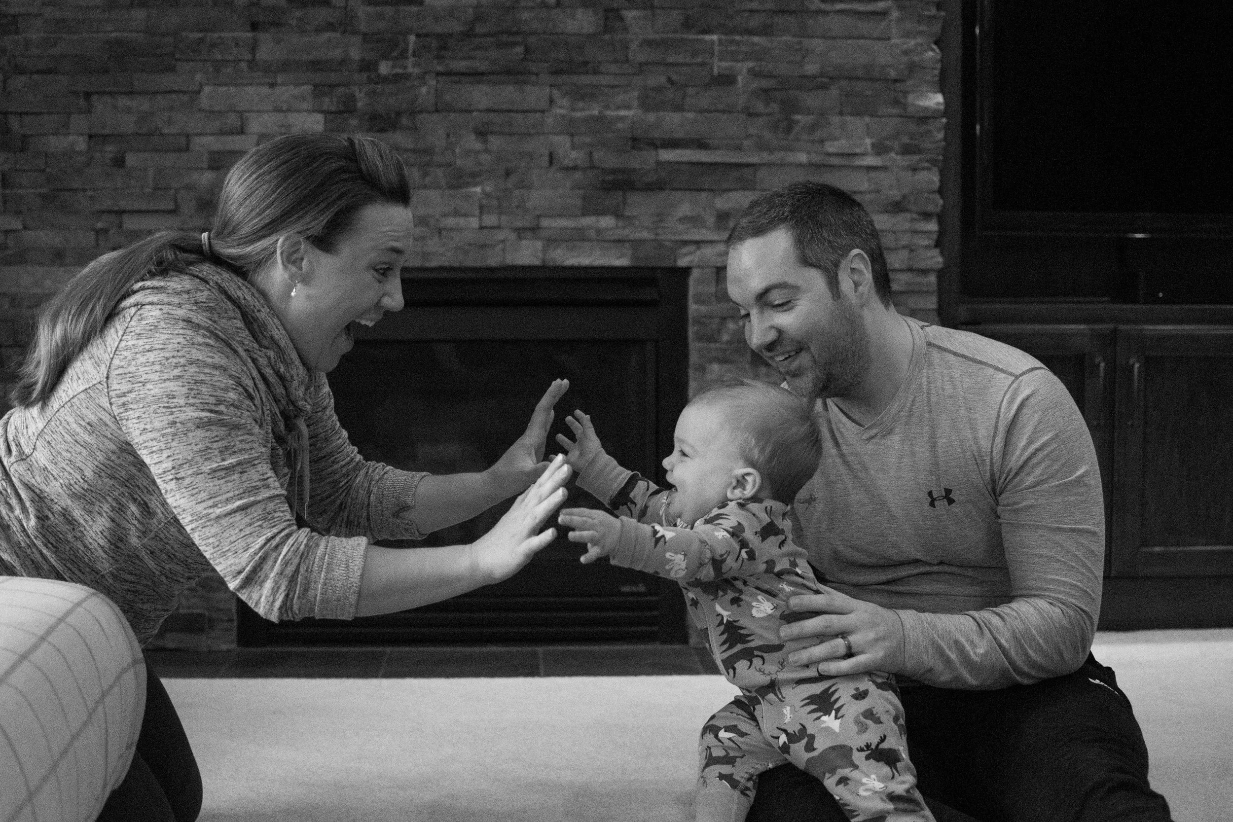 Mom holding out hands to young son, dad holding son on his lap in living room during Appleton Photo Session