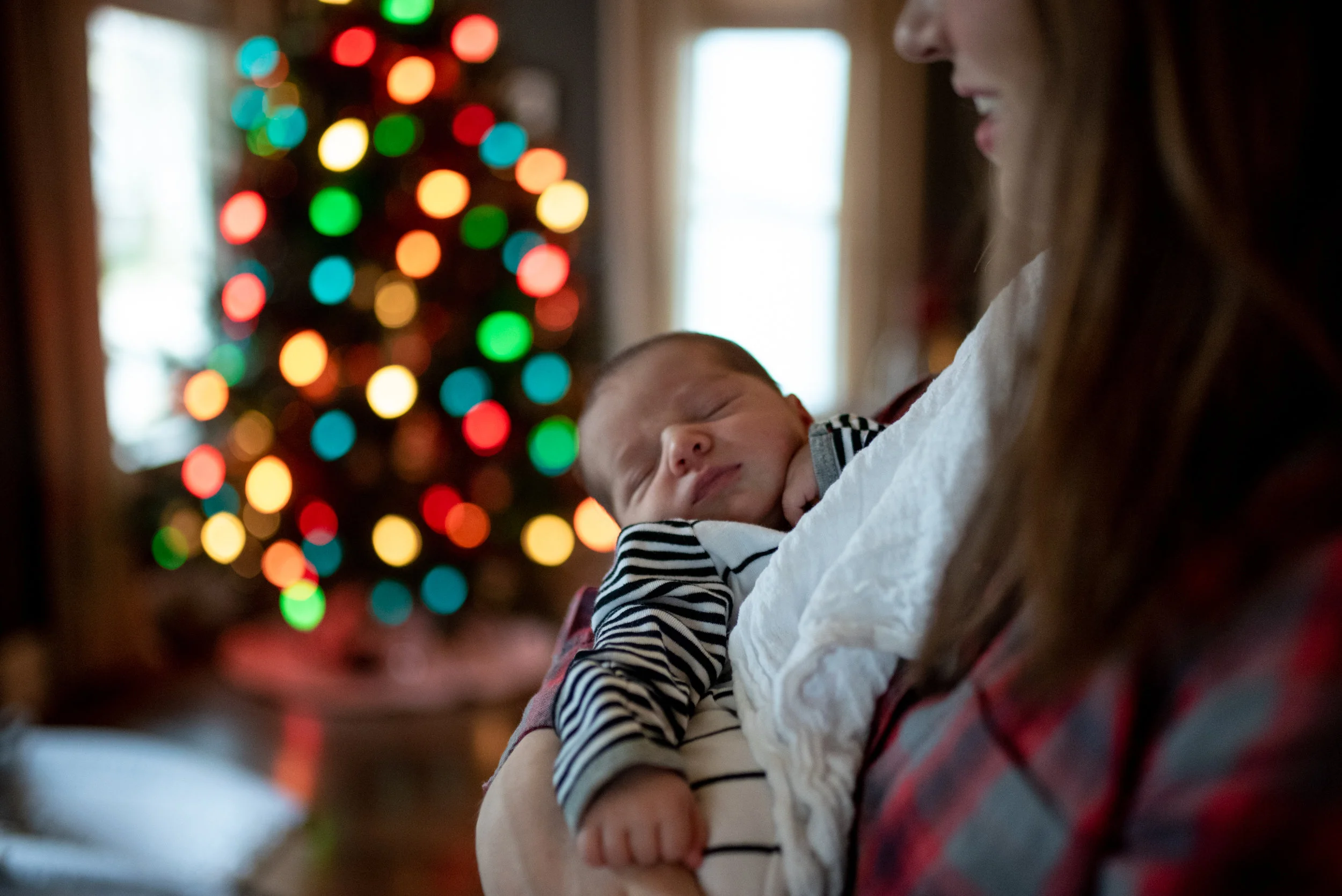 Mom holding newborn baby in her arms in front of a colorful Christmas tree
