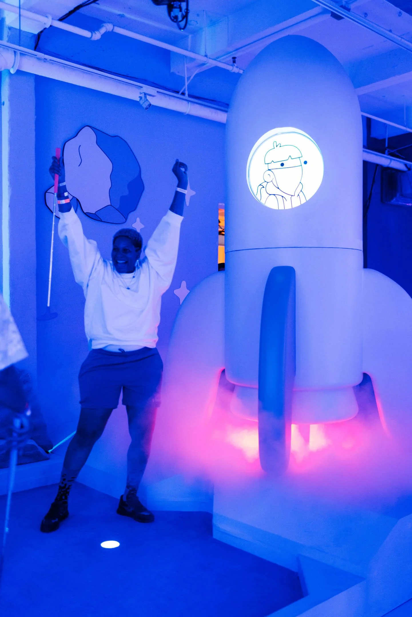 A woman with short hair, wearing a white sweatshirt and dark shorts, smiling and raising her arms in excitement next to a large, stylized rocket sculpture illuminated with pink and blue lights. The background features a wall with space-themed artwork
