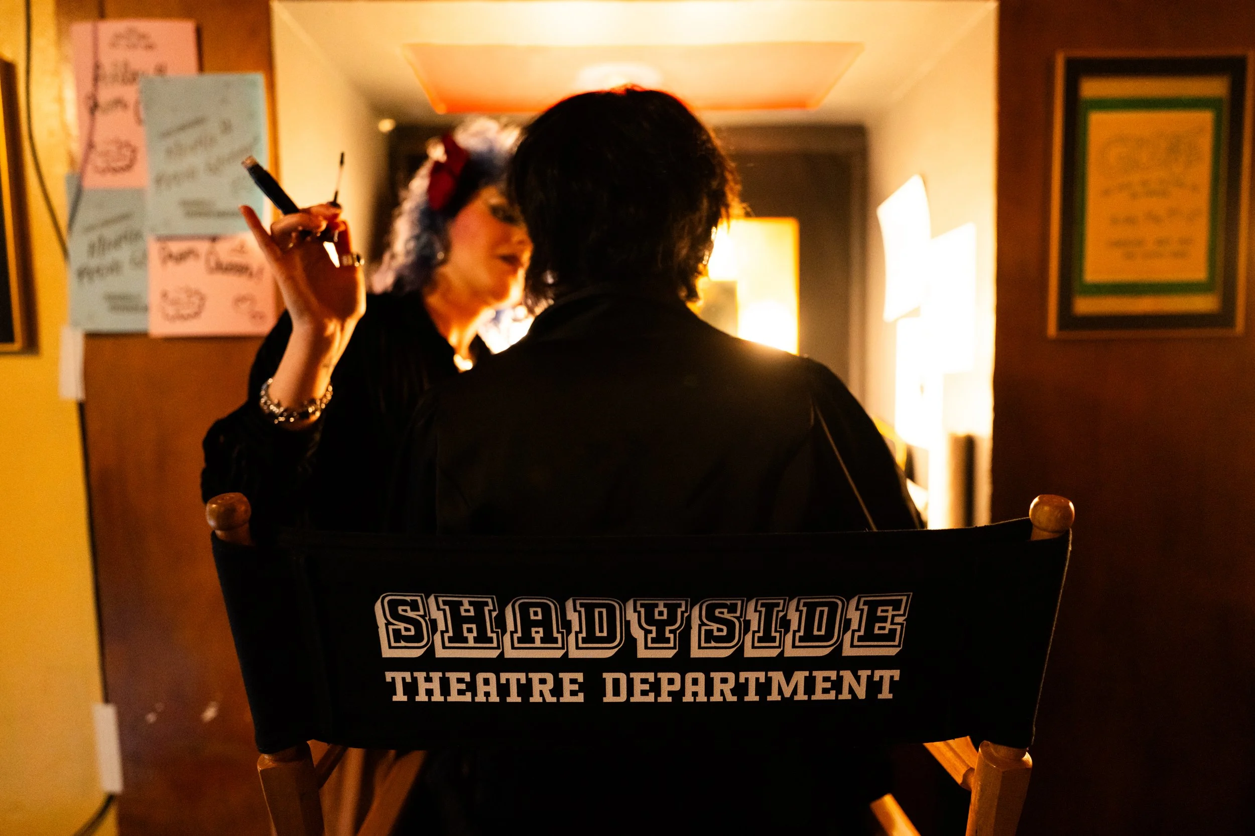 Two people are sitting and conversing in a dimly lit room. One person's back is facing the camera, and the other is holding a cigarette and appears to be speaking. The chair they are sitting on has 'Shady Side Theatre Department' written on the back.