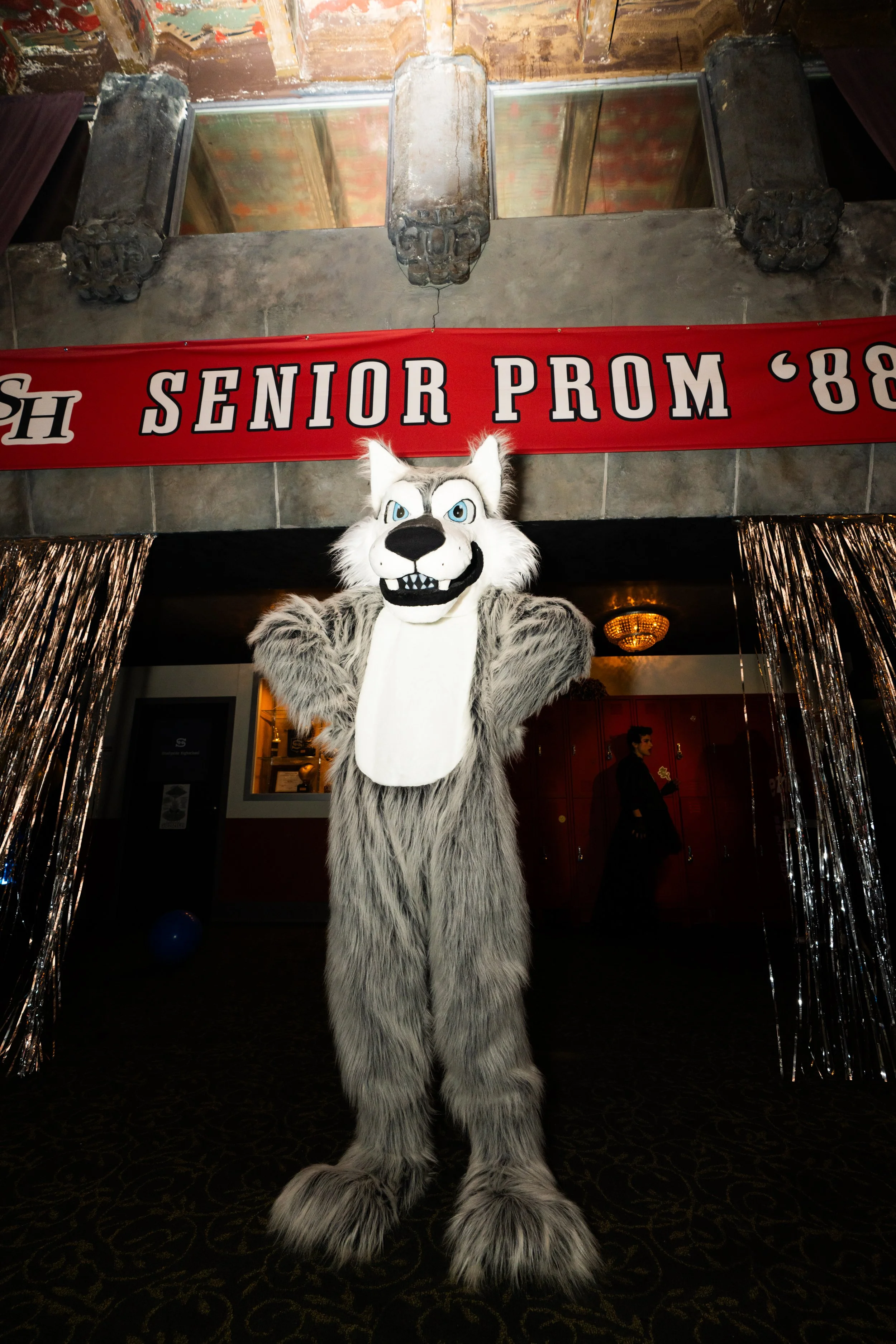 A person in a large wolf mascot costume standing in front of a red banner that reads 'Senior Prom '88' at a party or event, with metallic streamers on the sides and a background of an indoor setting.