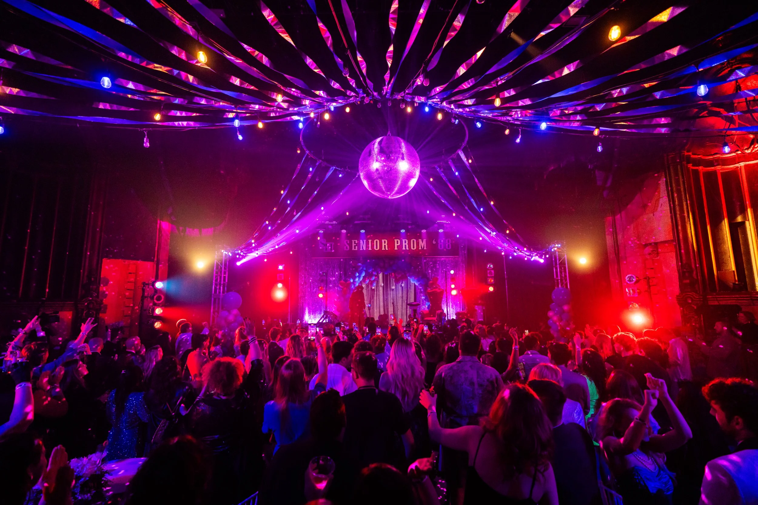 Crowd dancing at a prom party with colorful lights and a disco ball, decorated with streamers and balloons.