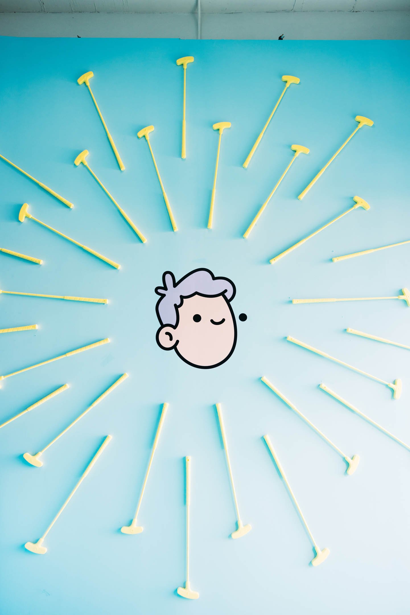 A wall decorated with 20 yellow ice cream swabs arranged in a circular pattern around a cartoon face of a person with gray hair, winking and smiling on a blue background.
