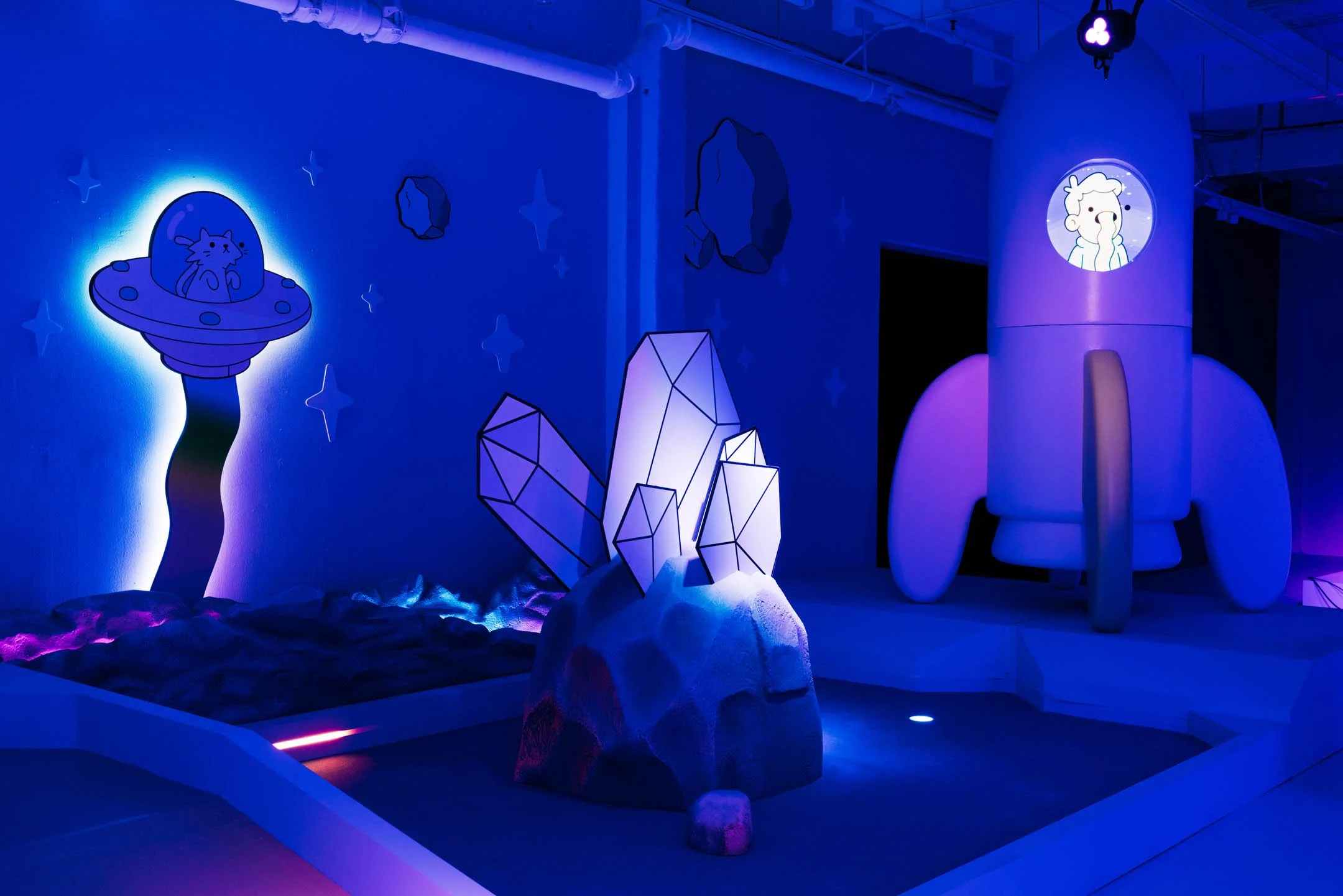 A space-themed indoor exhibit with glowing neon illustrations of a UFO with a cat inside, abstract geometric rocks with LED lights, and a cartoonish rocket with an animated character's face displayed on a porthole, all illuminated with blue and purpl