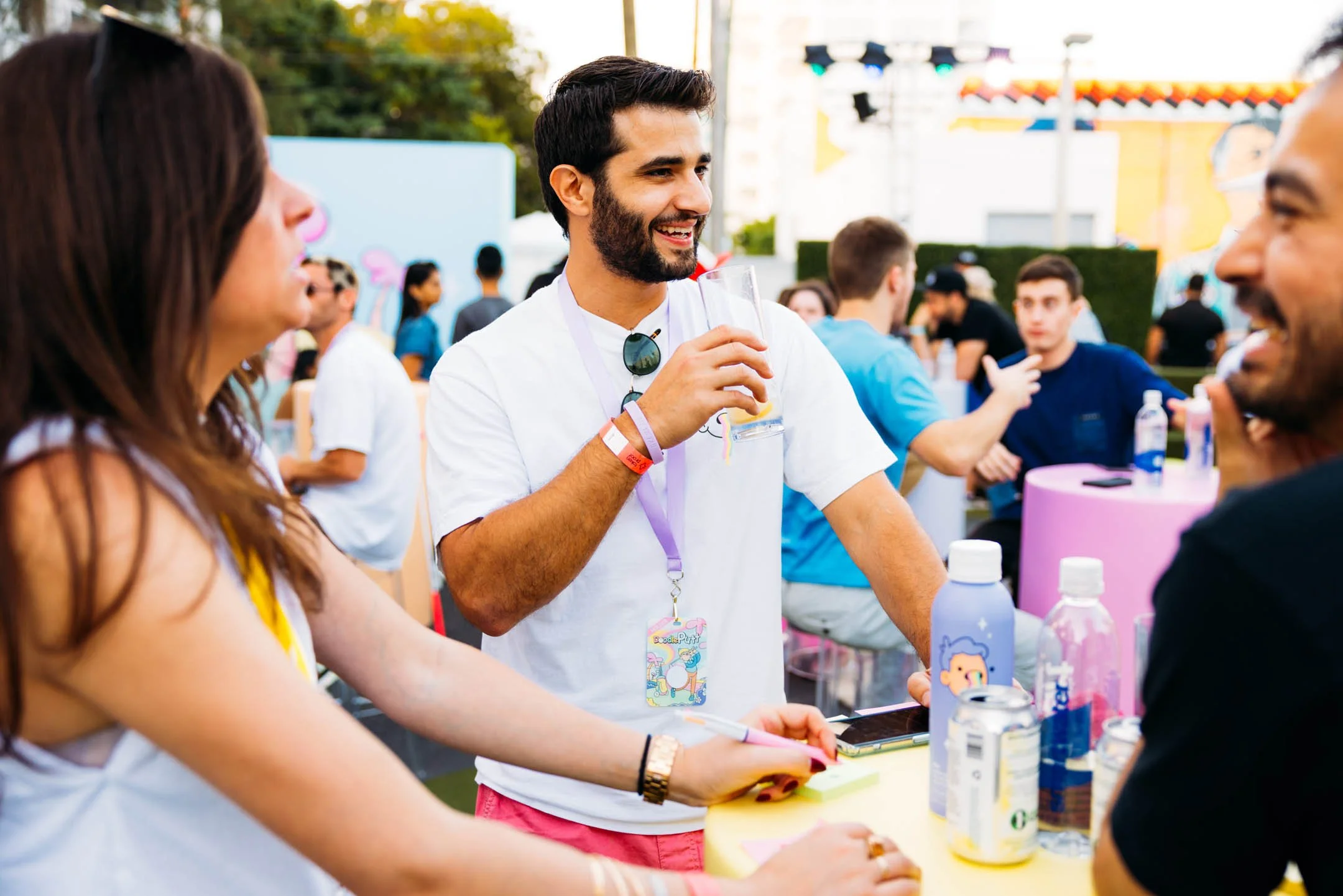 A young man holding a drink and laughing while engaging in conversation with two women at an outdoor event with colorful decorations and many people in the background.