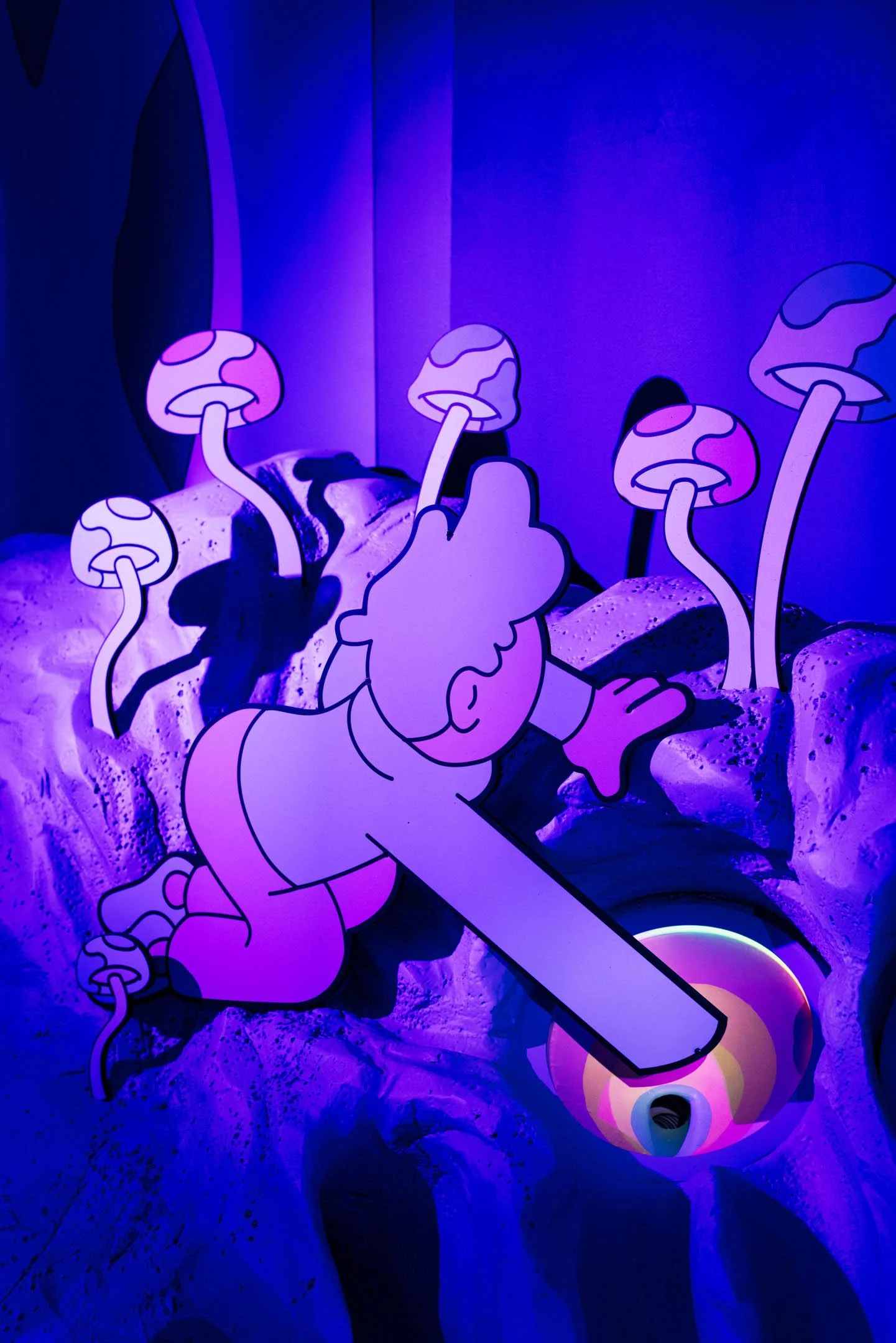 A neon art installation features a cartoon-style girl reaching toward a circular light with a yellow and pink glow, surrounded by stylized mushrooms emitting purple light, set against a textured blue and purple background.