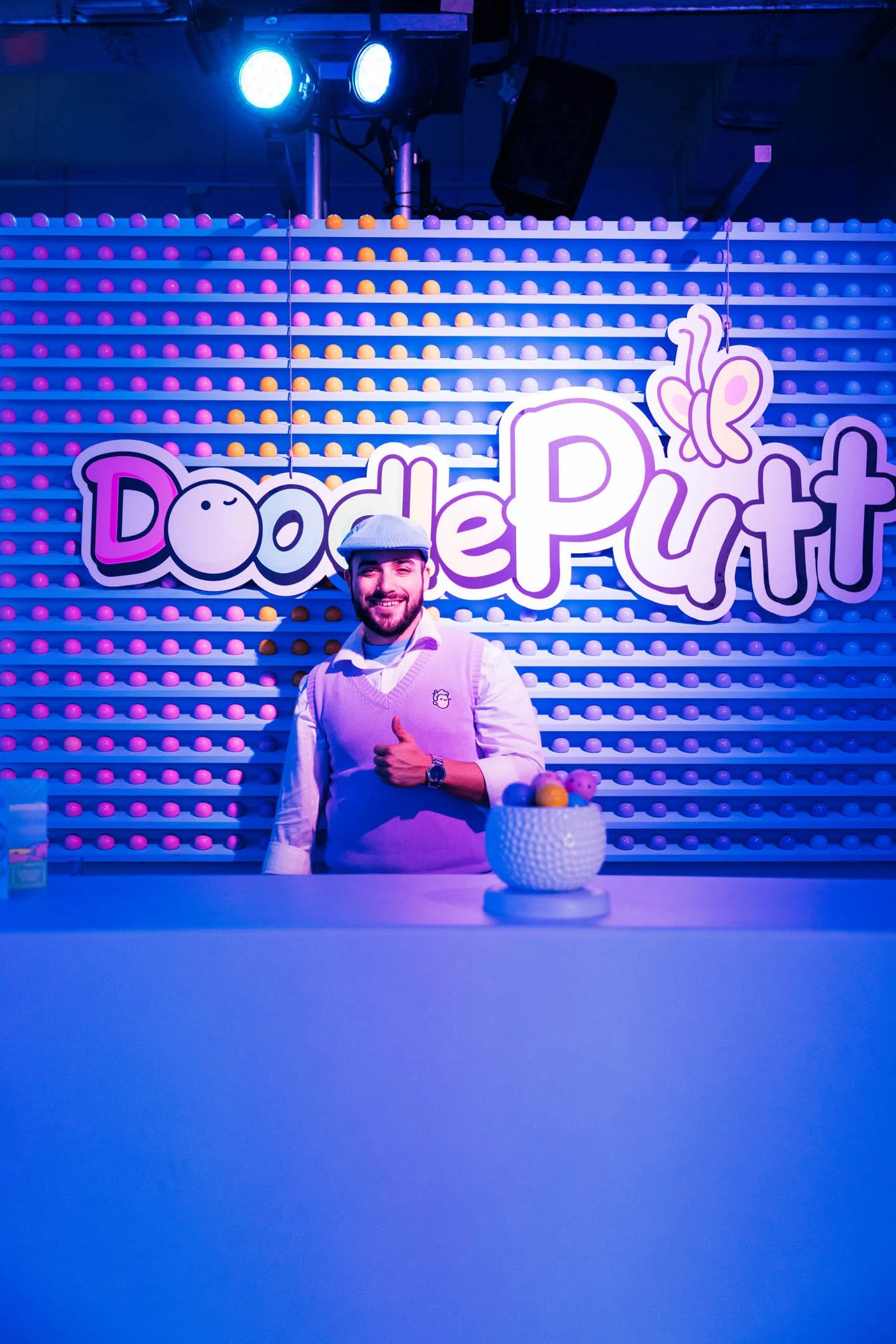 A man smiling and giving a thumbs-up in front of a colorful, playful sign that says 'DoodlePutt' with a butterfly graphic, at an indoor mini-golf or entertainment venue, illuminated with vibrant blue and purple lighting, with a table holding a bowl o