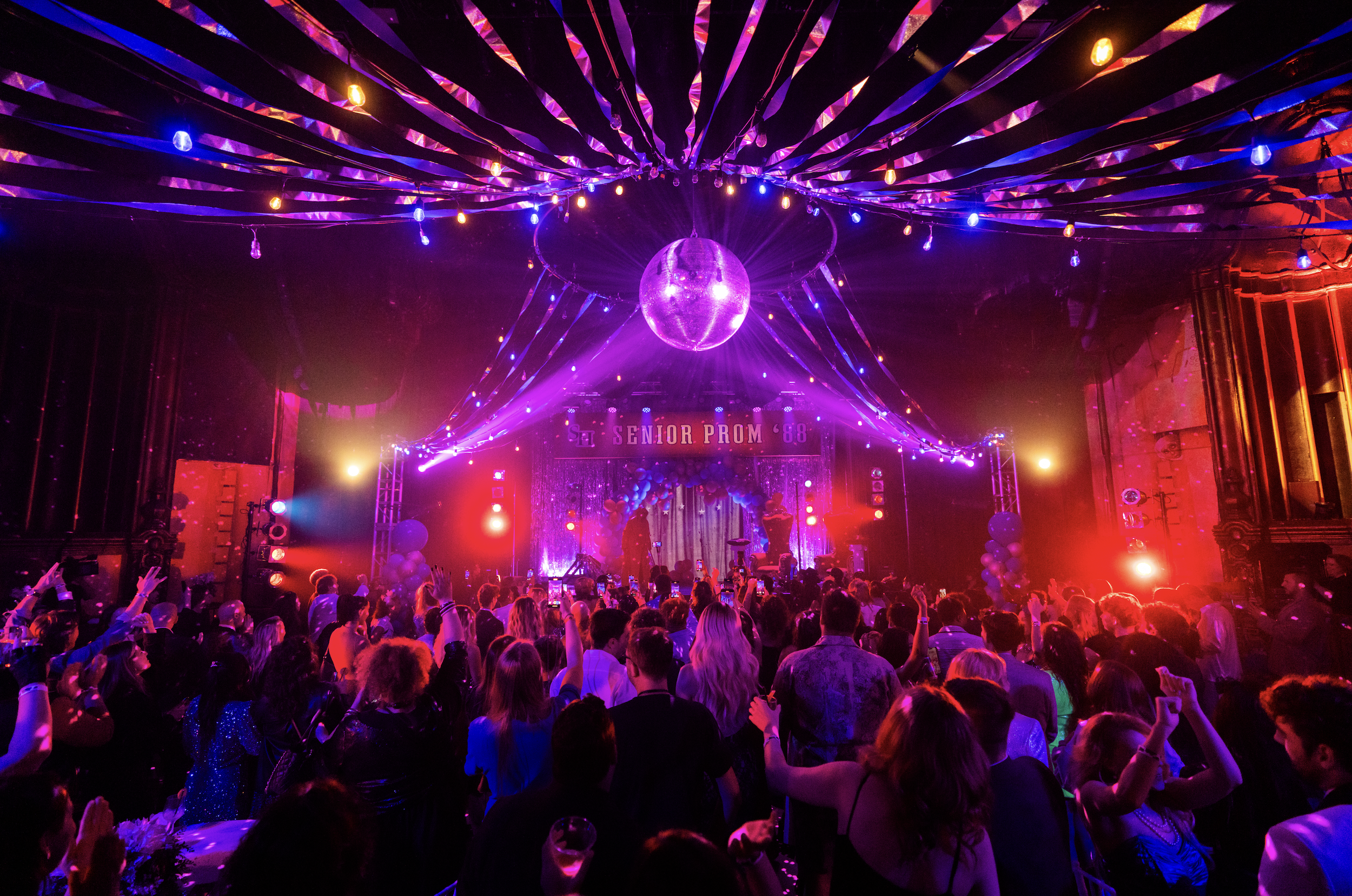 A lively prom party with colorful lights, a disco ball, and a crowd of young people dancing in a decorated venue with a stage. The sign reads 'Senior Prom '68'.