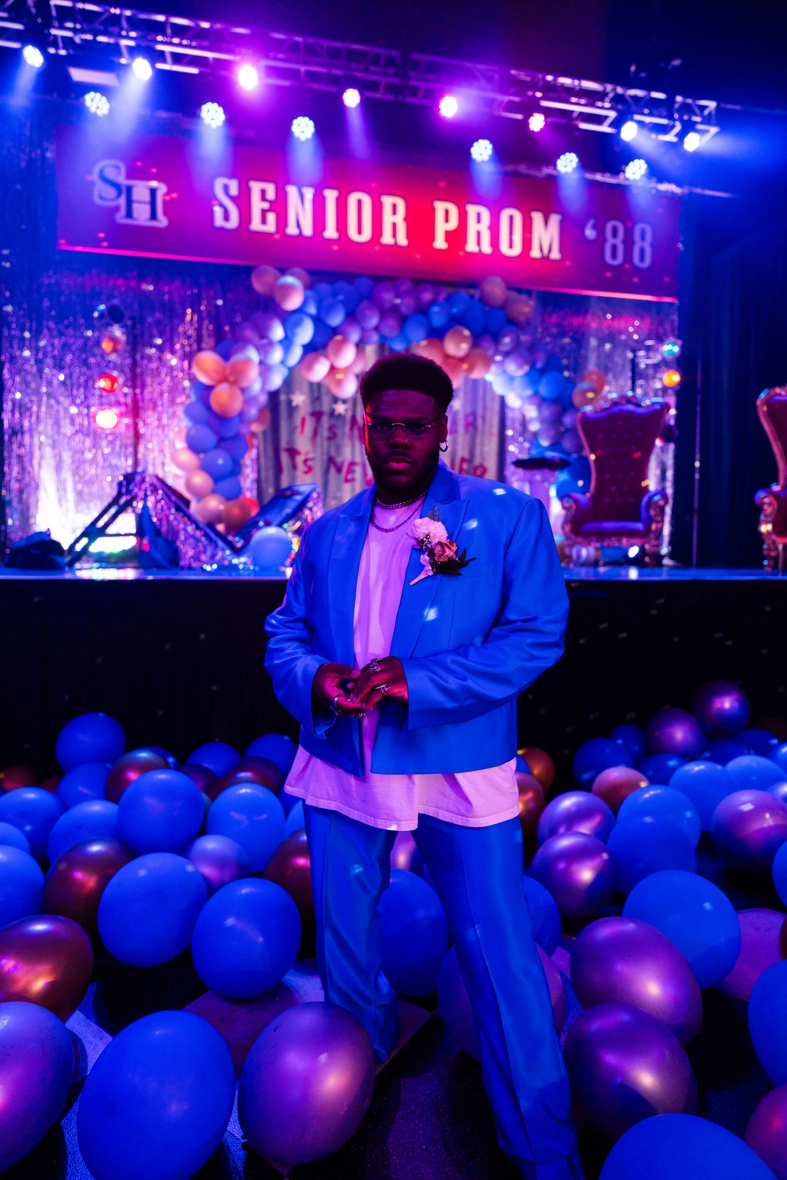 A person standing in front of a decorated stage at a senior prom, with balloons on the floor. The person is wearing a light blue suit with a white shirt, glasses, and jewelry. The stage has chairs, balloon arch, and a banner that reads 'Senior Prom '