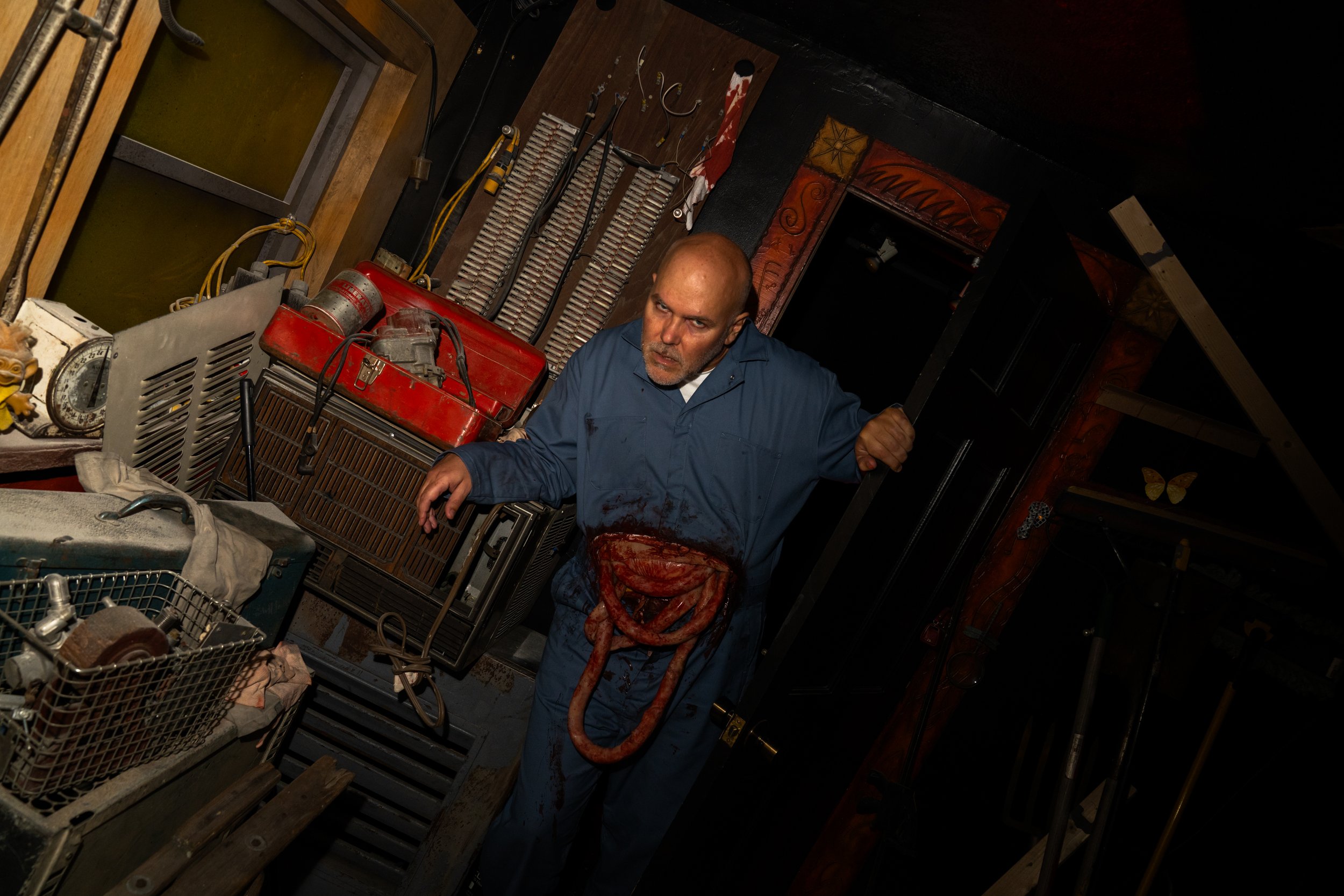 A man dressed as a mechanic or worker in a blue jumpsuit, with a bloody apron or stained belt, is standing in a cluttered workshop or garage filled with tools and equipment. The man has a serious or intense expression.