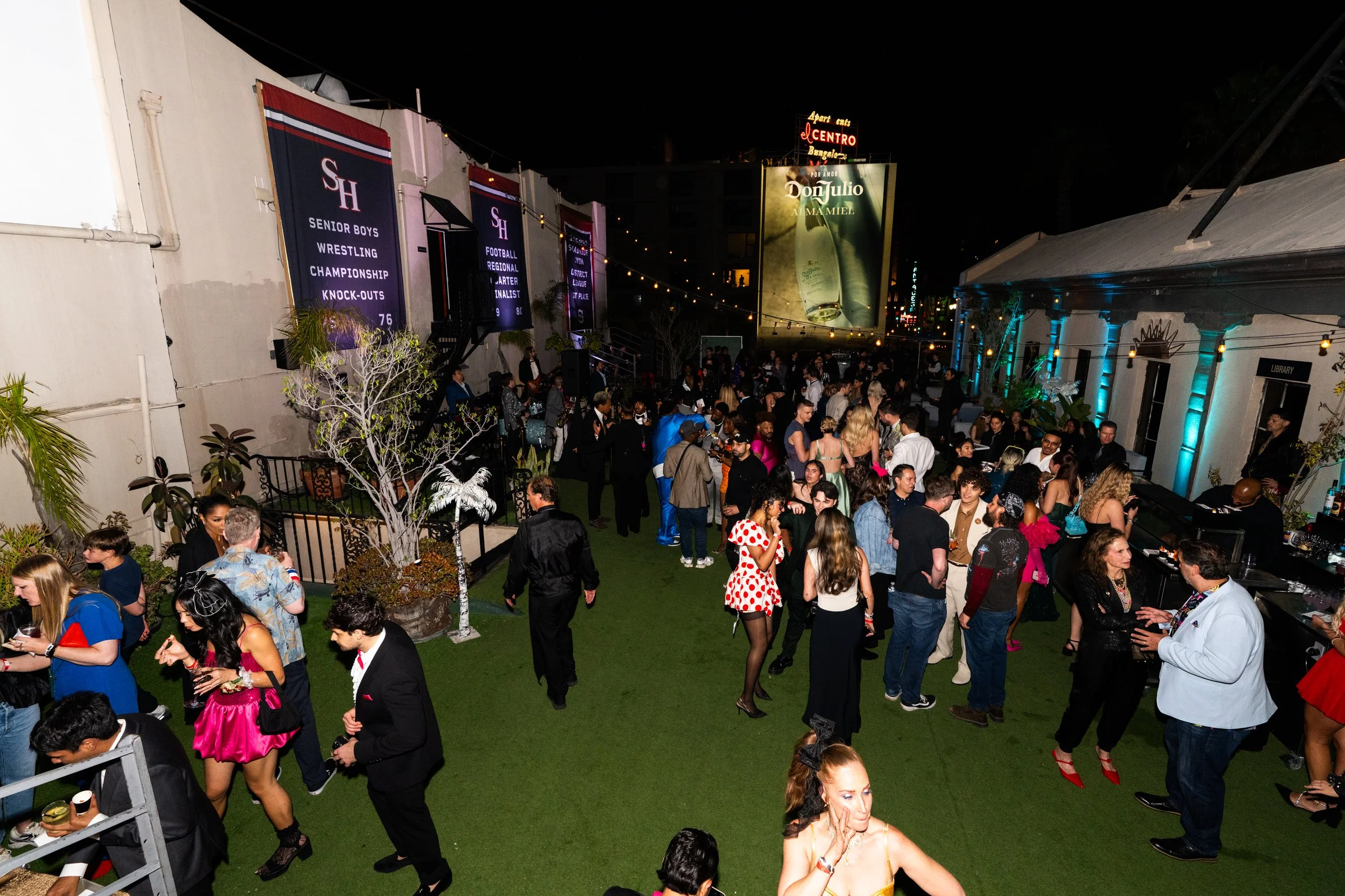 Crowd of people socializing at an outdoor party at night, with banners on the wall for high school wrestling championships, a large advertisement, and colorful lighting on a building.