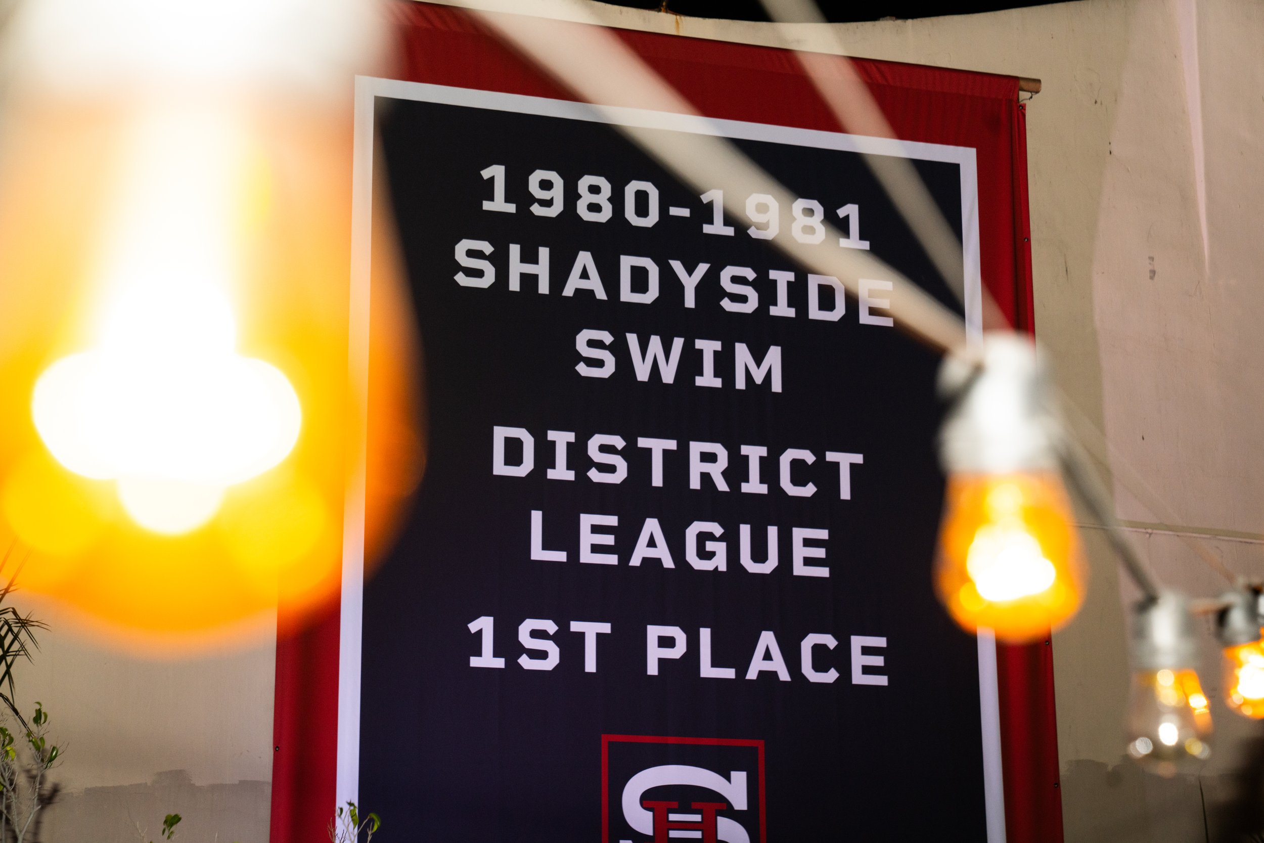 A black banner with white text reads '1980-1981 SHADYSIDE SWIM DISTRICT LEAGUE 1ST PLACE'. String lights are hanging in the foreground, partially obscuring the banner.