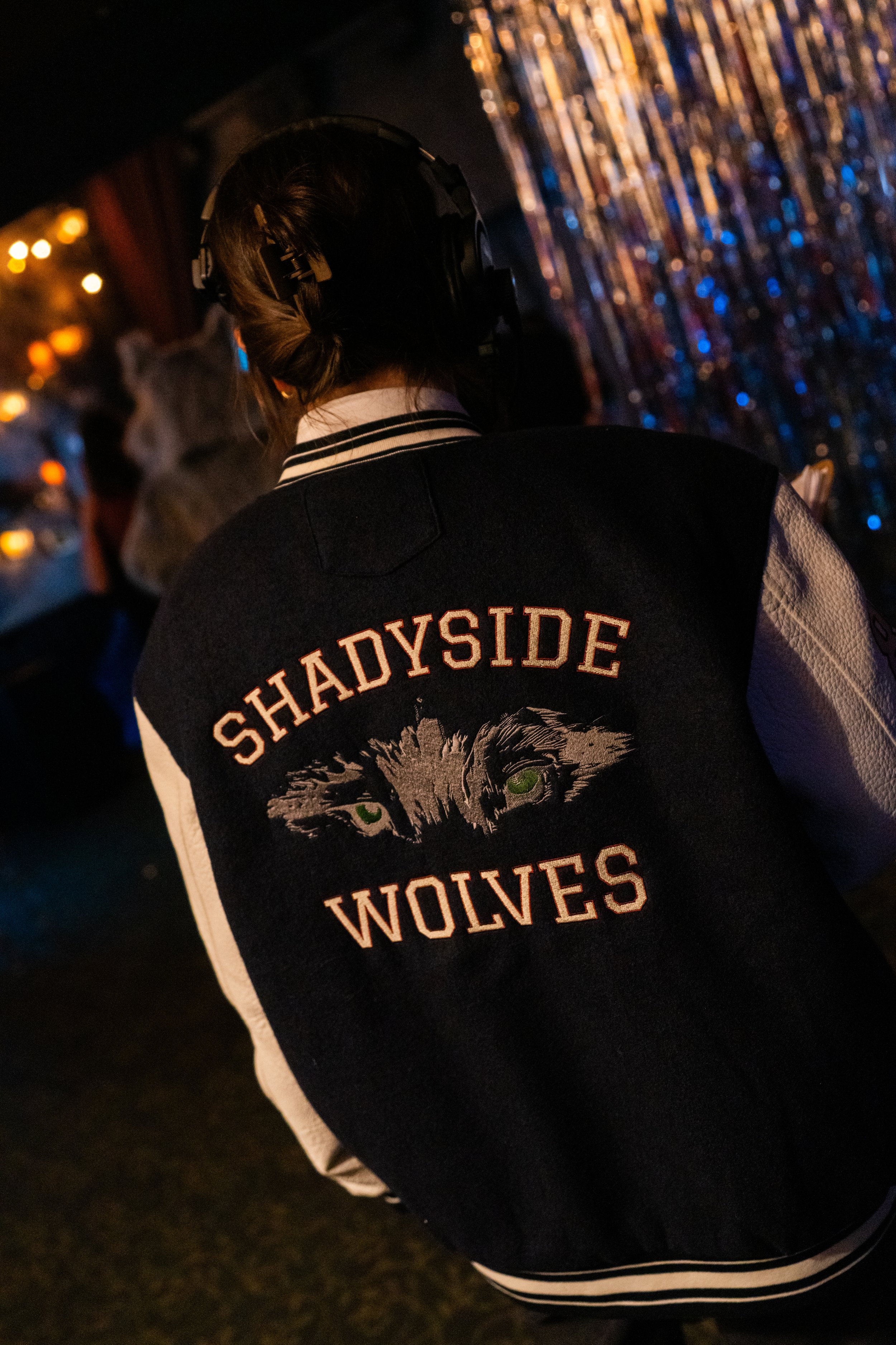 A person wearing a 'Shadyside Wolves' jacket with a wolf graphic, seen from the back, in a dimly lit setting with colorful hanging lights in the background.