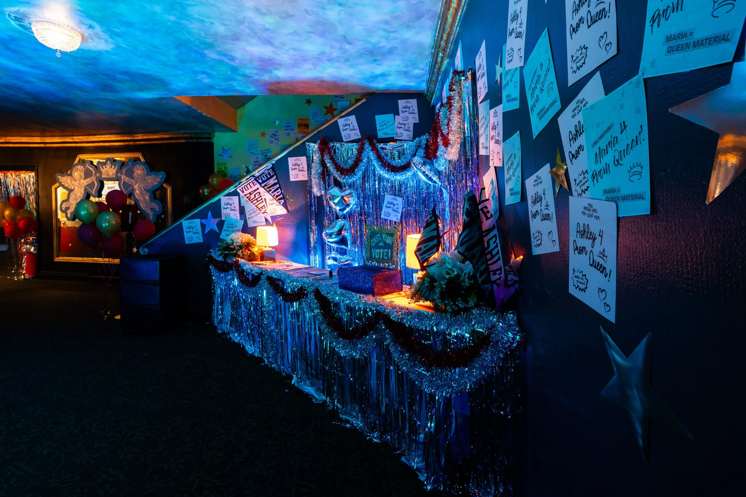 Decorated table with tinsel, flags, and flowers in a dimly lit room celebrating a pageant or event, with congratulatory messages and ballots on the wall.