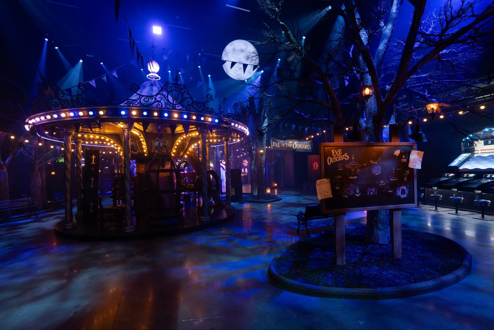 Empty indoor amusement park or arcade area with a carousel ride, themed for Halloween, decorated with a large moon and eerie trees, illuminated with purple and orange lights, and a sign for 'Eve of the Outcasts'.
