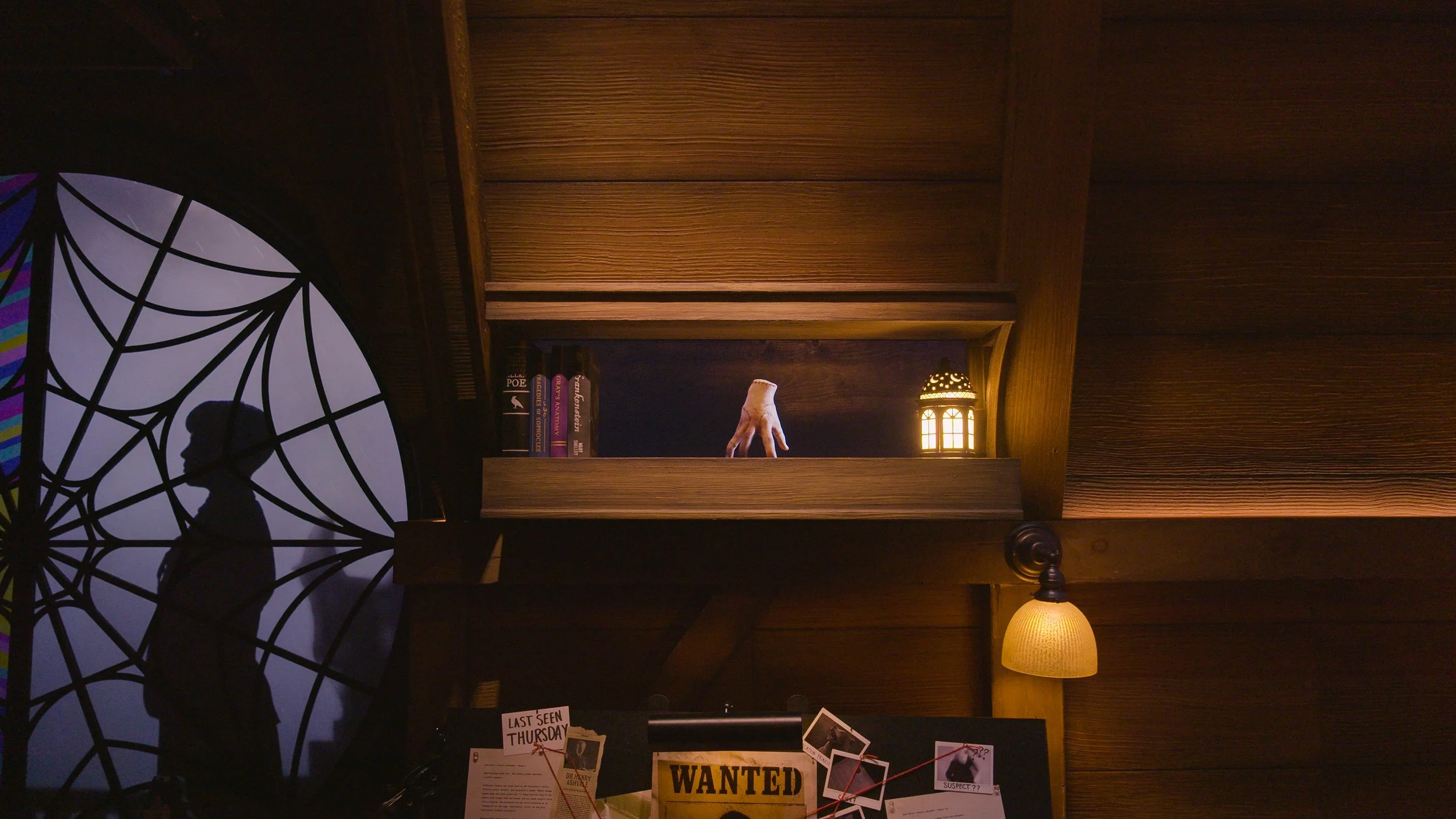 A silhouette of a man with a hat and briefcase, standing in front of a decorative stained glass window with a spider web pattern, inside a dimly lit room with wooden walls, a bookshelf, a small lamp, and a bulletin board with photos and notes.