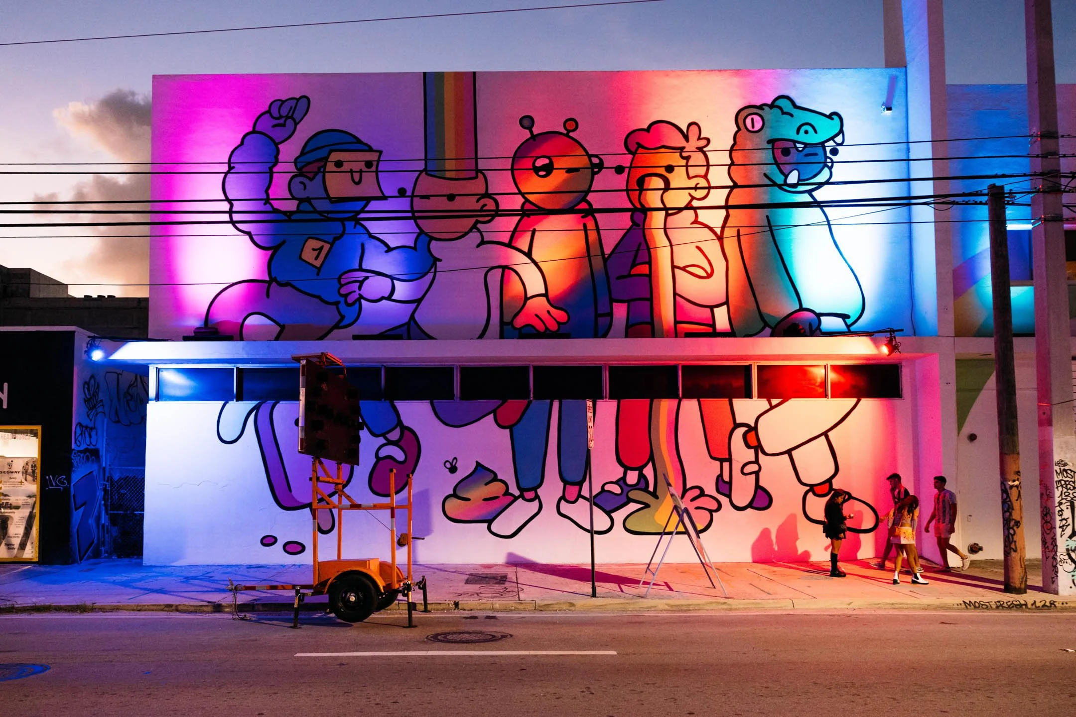 Street mural of cartoon characters with rainbow accents, illuminated by colorful lights at dusk, with three people walking by.