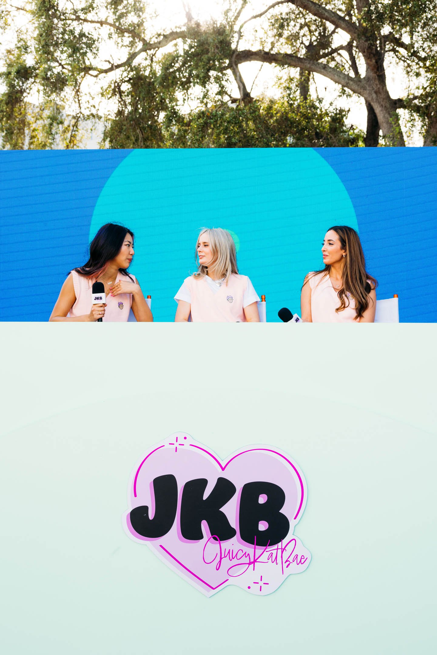 Three women seated at a table during an outdoor event, with a blue background and trees overhead. The women are engaging in a discussion, each holding a microphone, and are wearing light-colored tops. In front of a plain wall, there is a heart-shaped