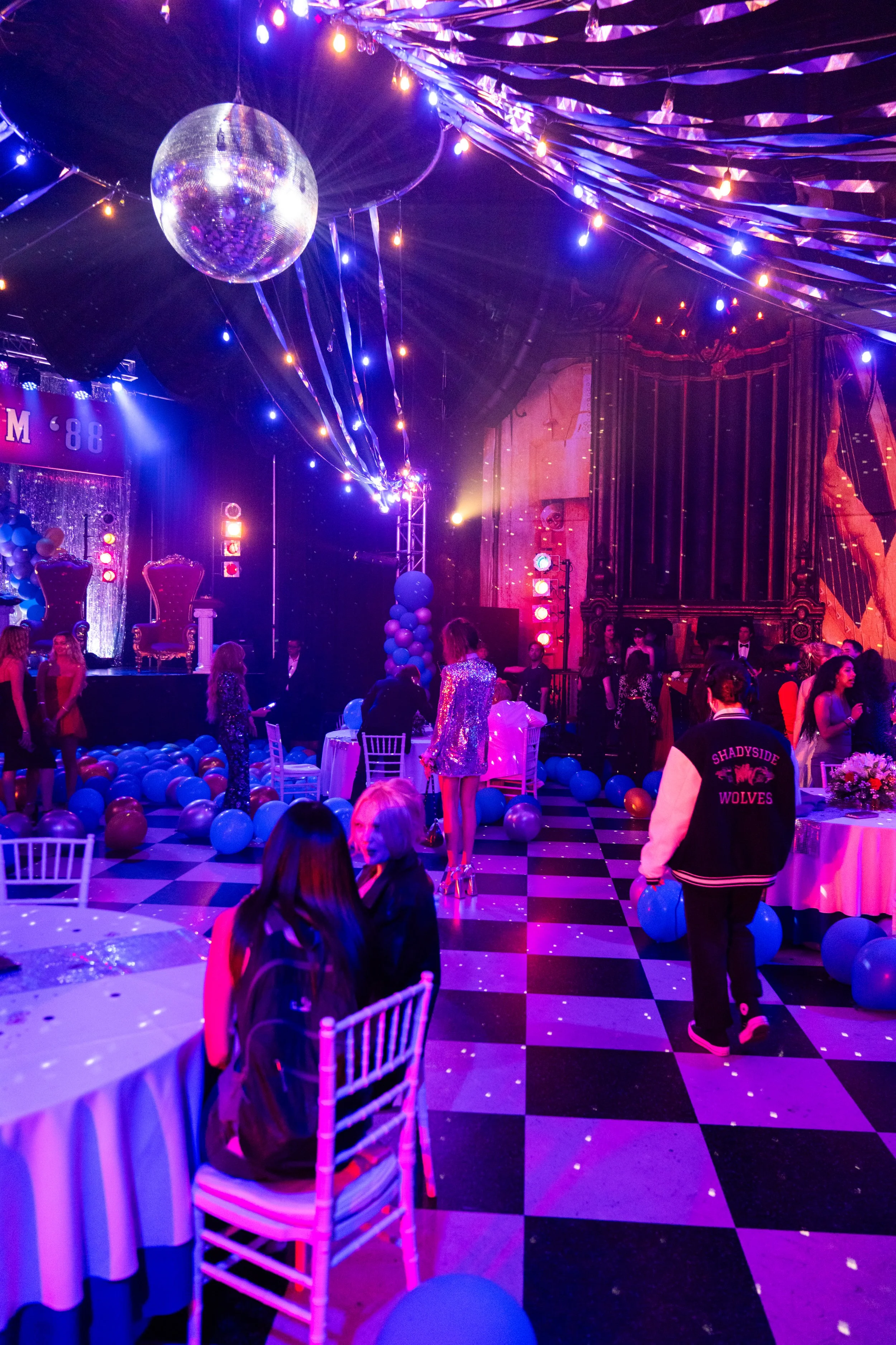 A lively party scene with colorful lighting, balloons, and a disco ball. People are socializing and dancing in a decorated venue with checkered flooring and a stage.