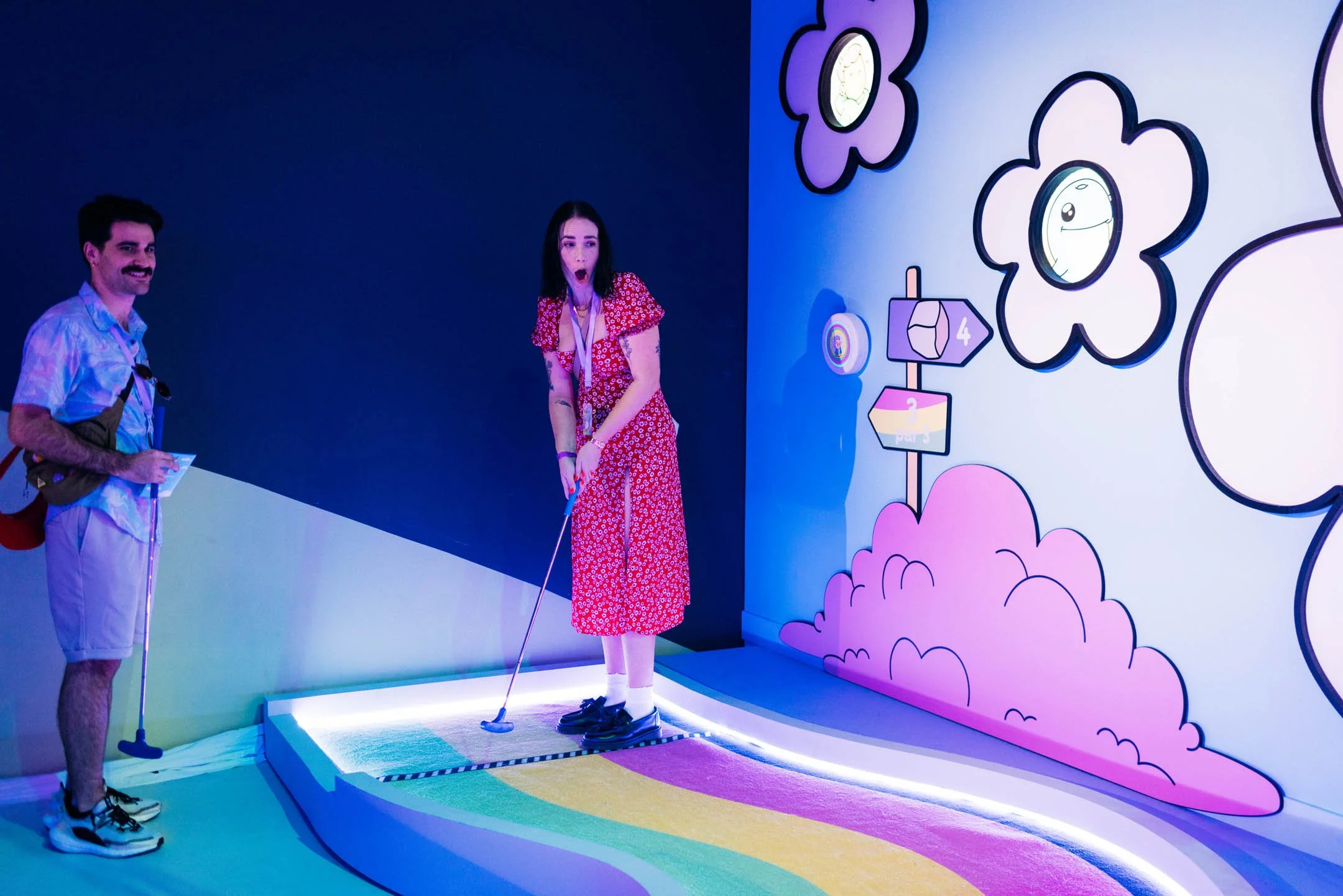 A woman in a red floral dress is riding a mini golf putt putt course with a man standing nearby. The scene is indoors with colorful, cartoon-style wall art including clouds and signs.
