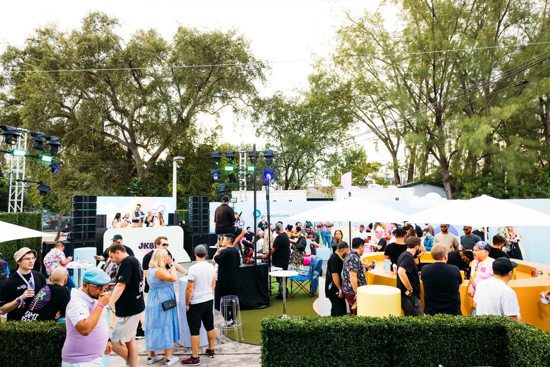 Outdoor music event with a stage, DJ, and crowds of people mingling around tables and in front of the stage, under umbrellas and trees.
